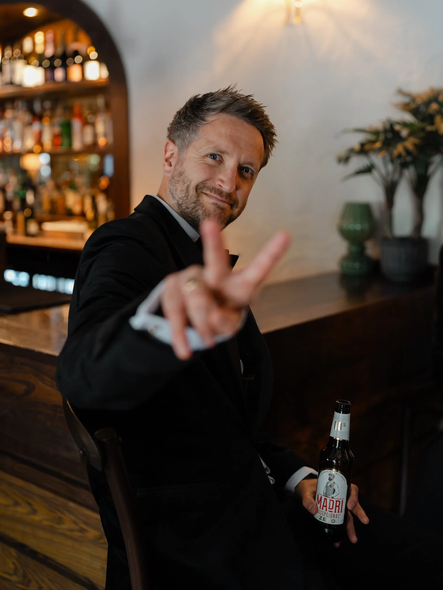 A man dressed in formal attire is sitting at a bar, holding a brown beer bottle with a white label that reads "Madri." He is smiling, making eye contact, and showing a peace sign with his right hand. The background shows a bar with various bottles on