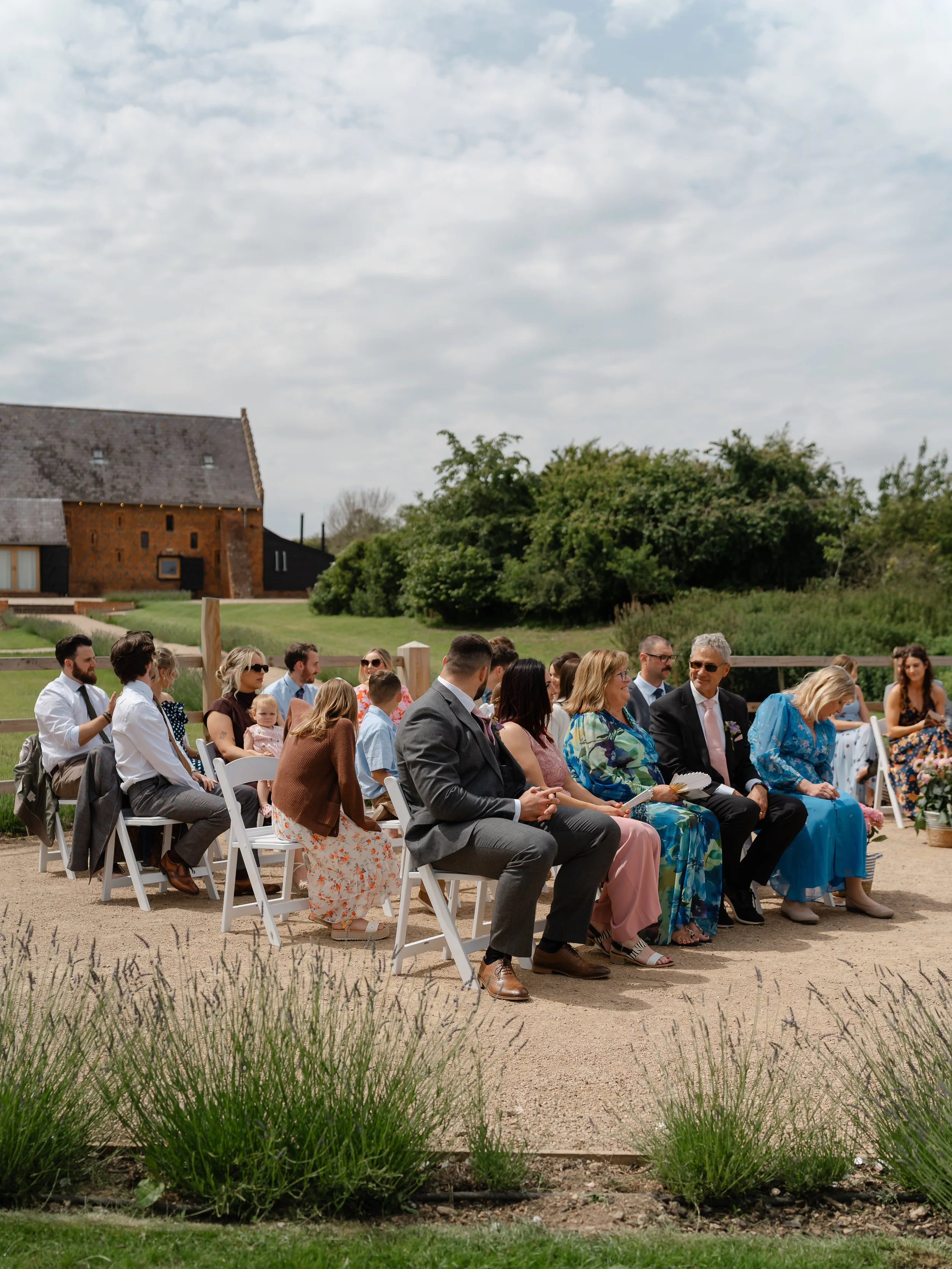 People sitting on white chairs outdoors during a daytime event, with a rural setting, a barn, trees, and a partly cloudy sky in the background.