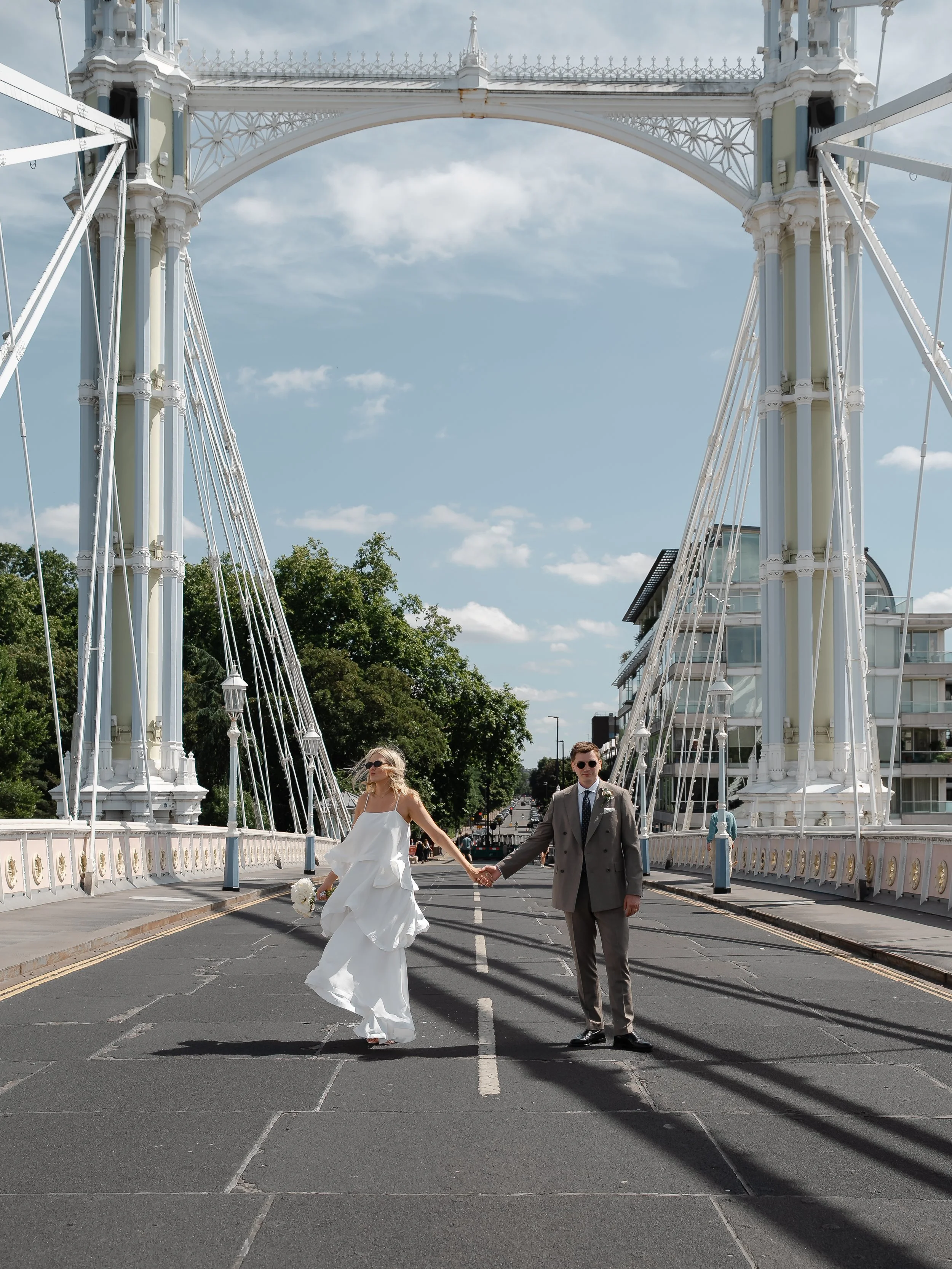 A bride and groom holding hands and walking across a bridge on a sunny day, with a background of trees and modern buildings.