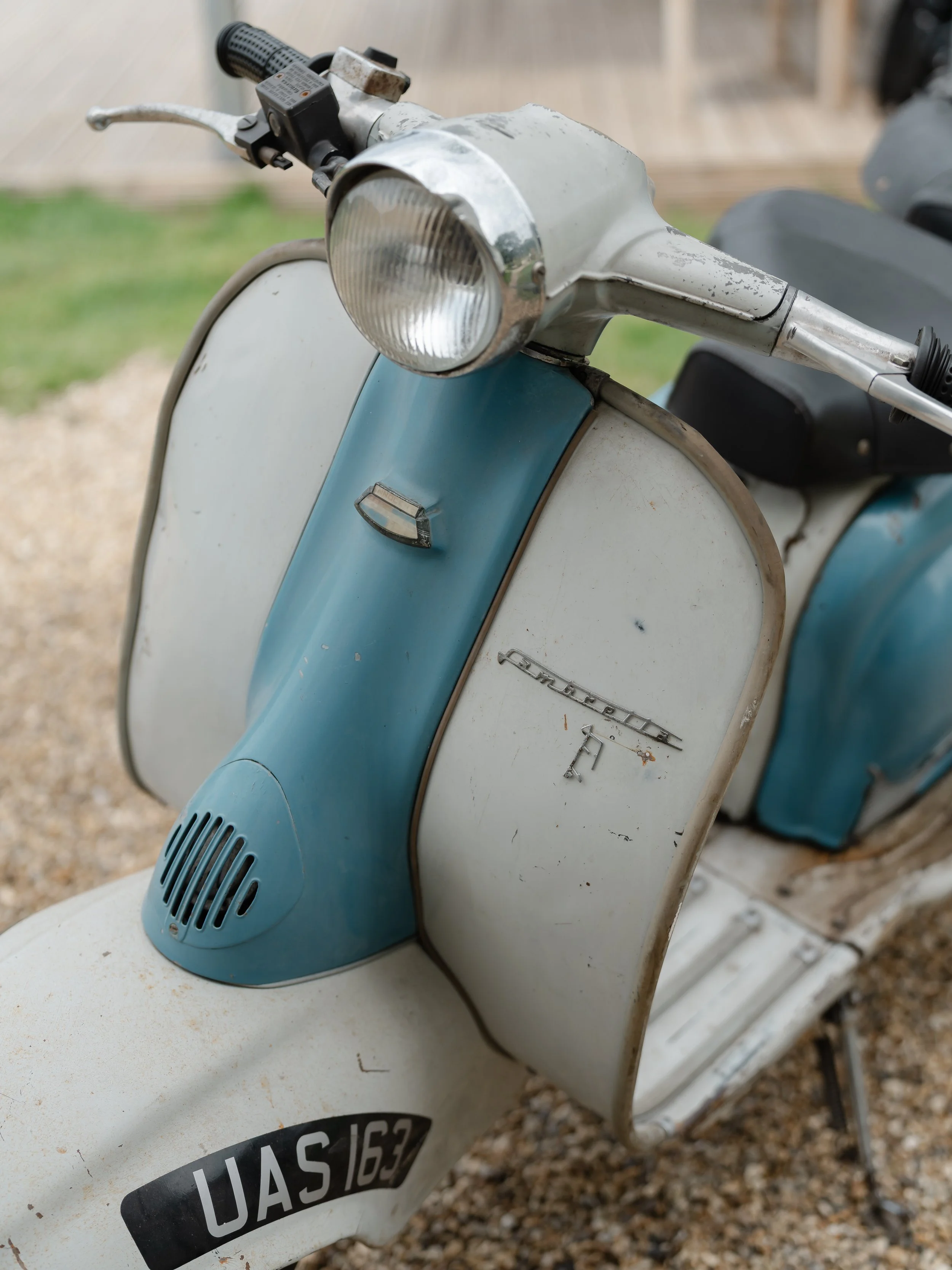 Close-up of a vintage blue and white Scootergirl scooter with a worn headlight, faded paint, and a license plate reading 'UAS 163', parked on gravel.