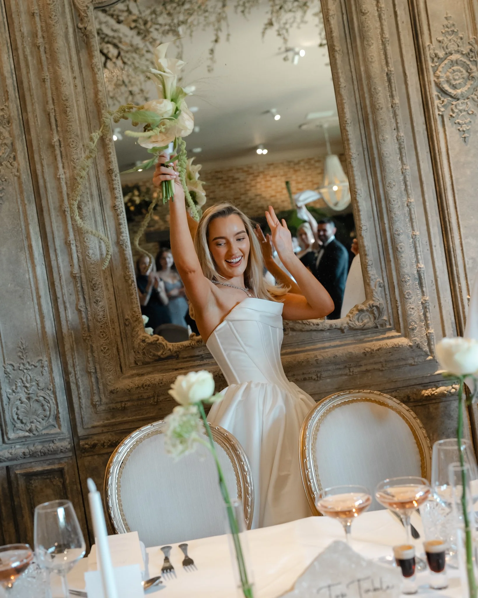 A smiling woman in a strapless white wedding dress holding a bouquet of white flowers, reflected in an ornate large mirror, with a wedding reception setting visible in the background.