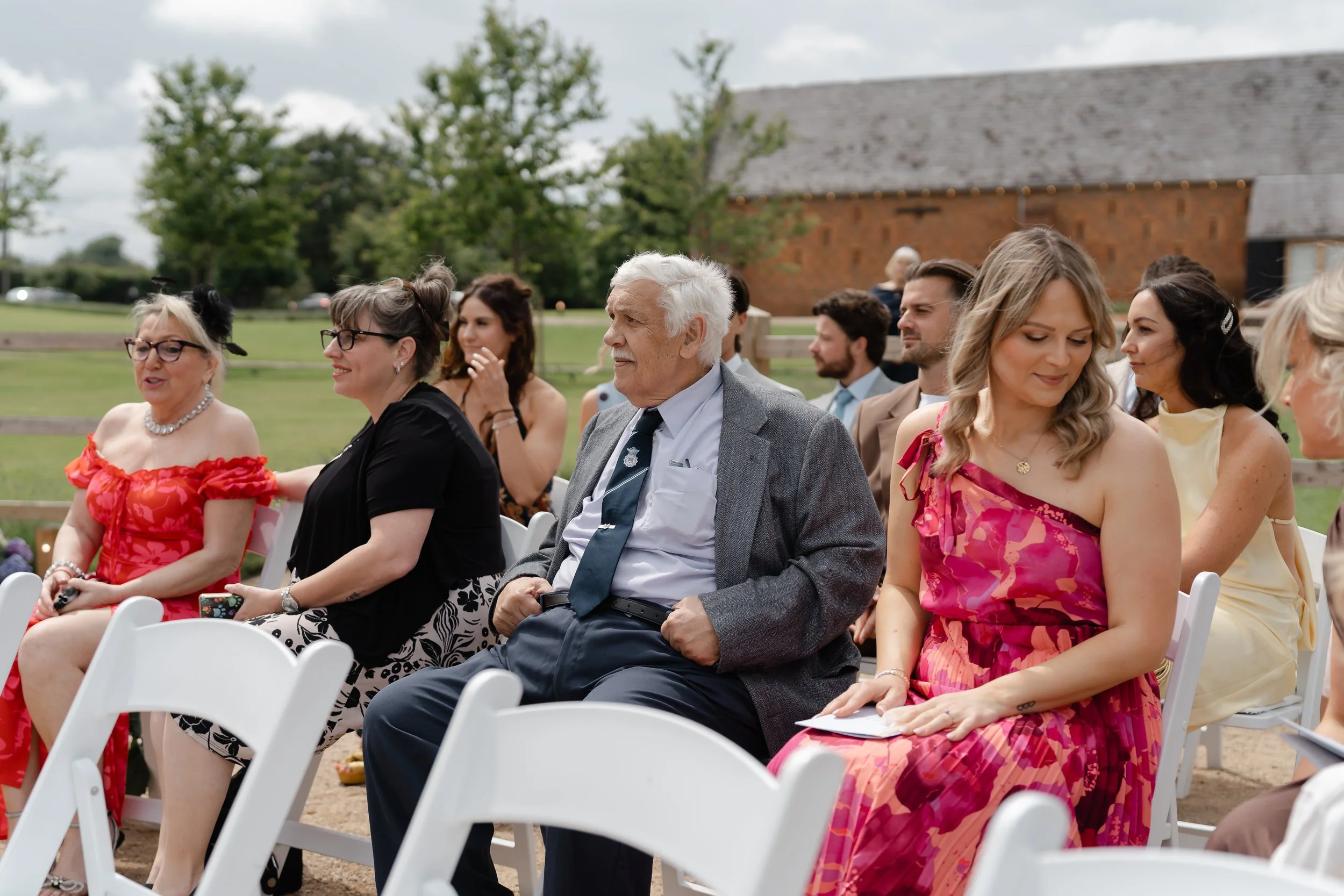 Group of people sitting outdoors at an event, with a brick building and green trees in the background. at Copdock Hall, Suffolk