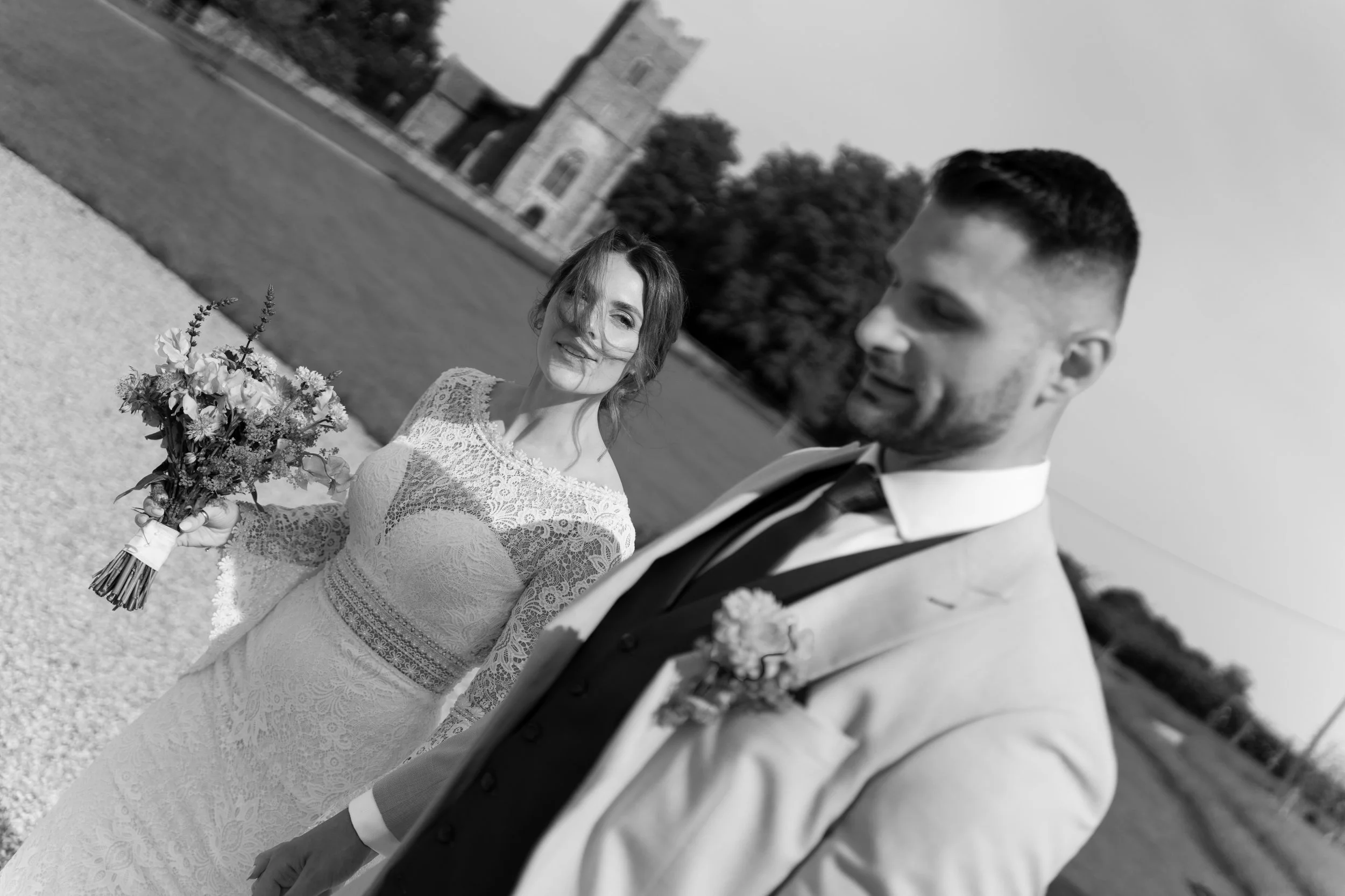 A black and white photo of a bride and groom outdoors, with a church building in the background. The bride is smiling and holding a bouquet, wearing a lace wedding dress. The groom is dressed in a suit with a tie, looking at the bride.
