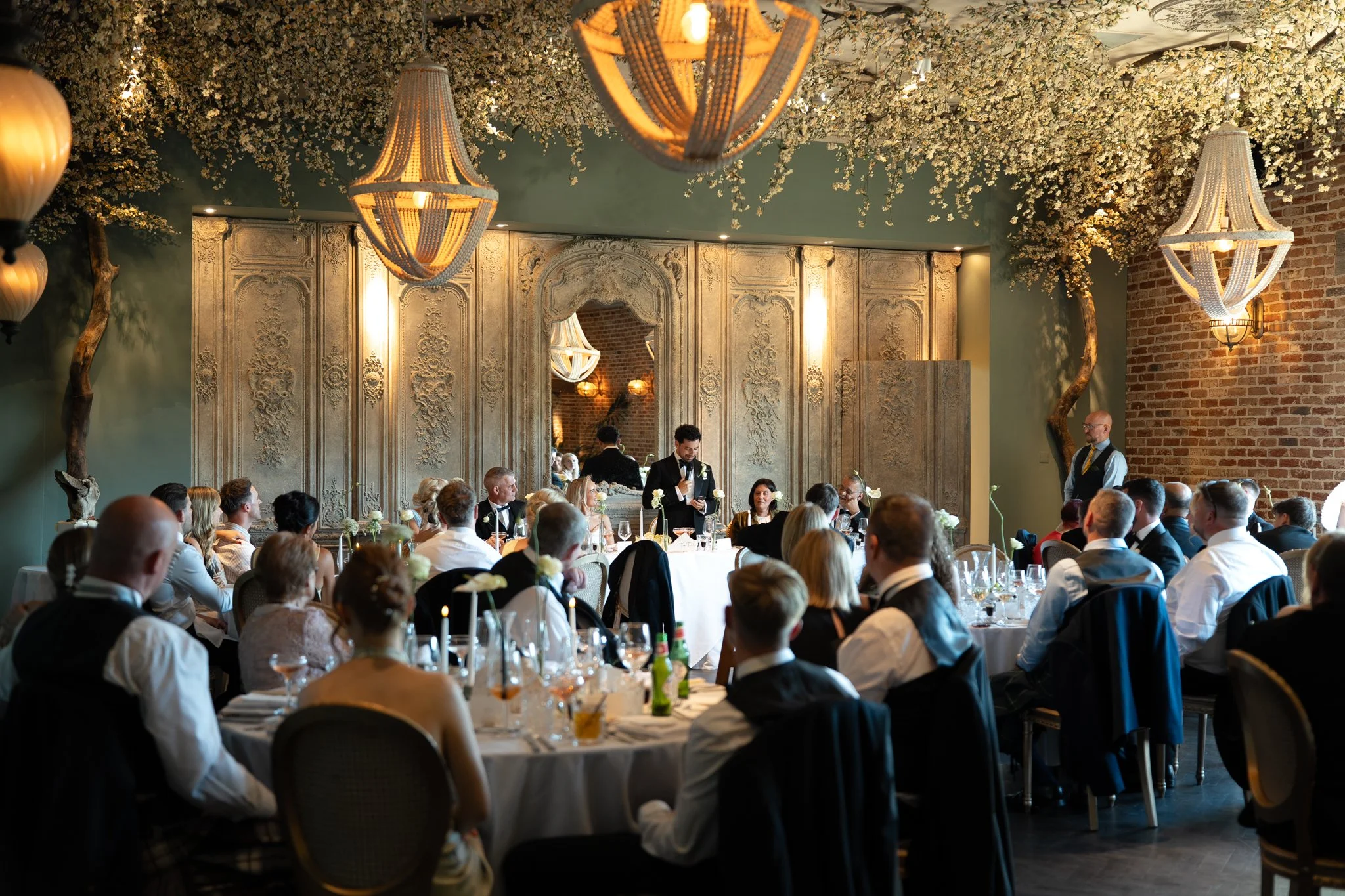A formal indoor event with many people seated at round tables decorated with white flower arrangements. A man is speaking at a microphone while others listen attentively. The ceiling is decorated with hanging chandeliers and abundant white floral arrangements.