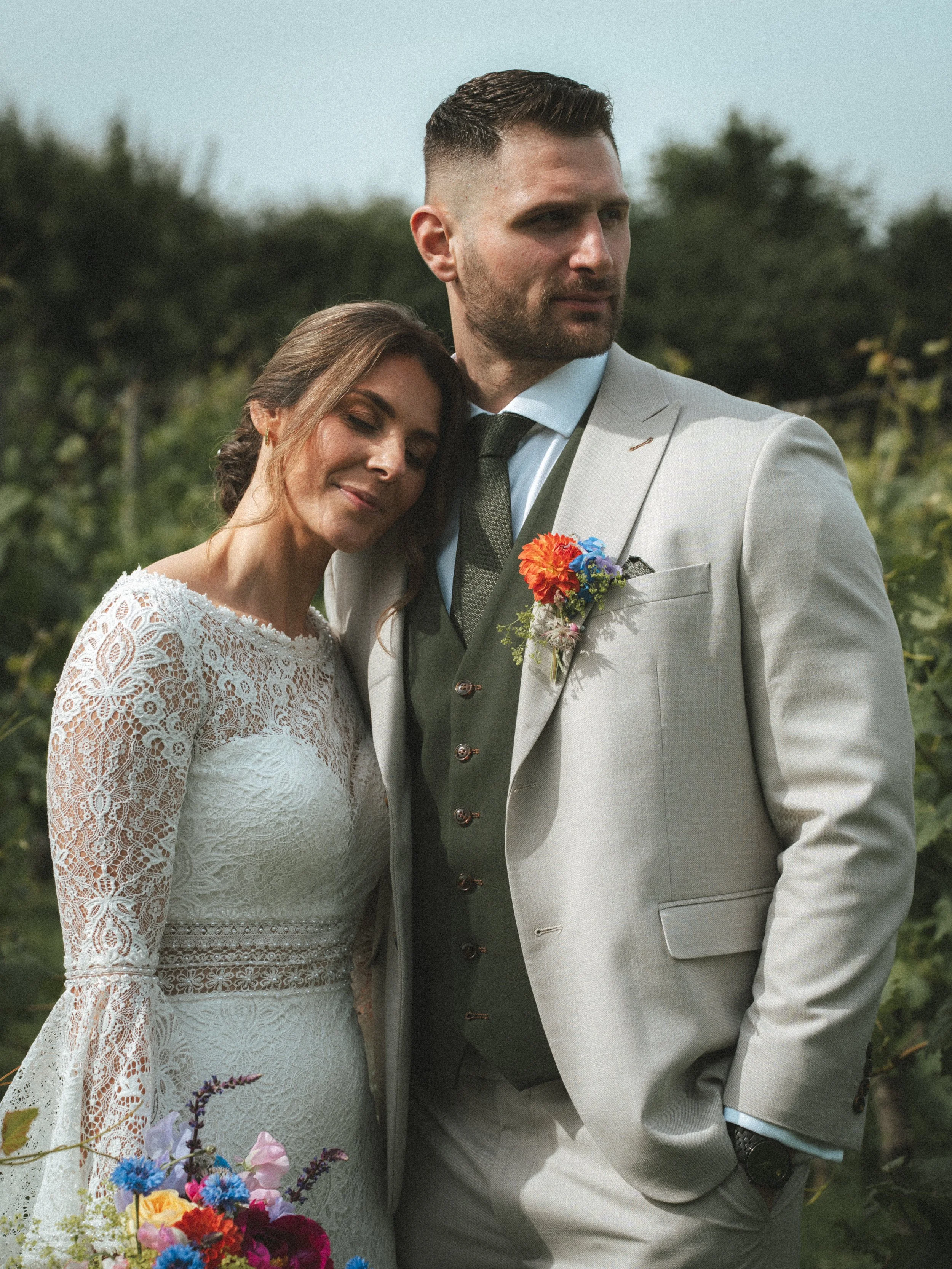 A bride and groom standing close outdoors during their wedding, with the bride resting her head on the groom's shoulder, both smiling gently, with trees in the background.