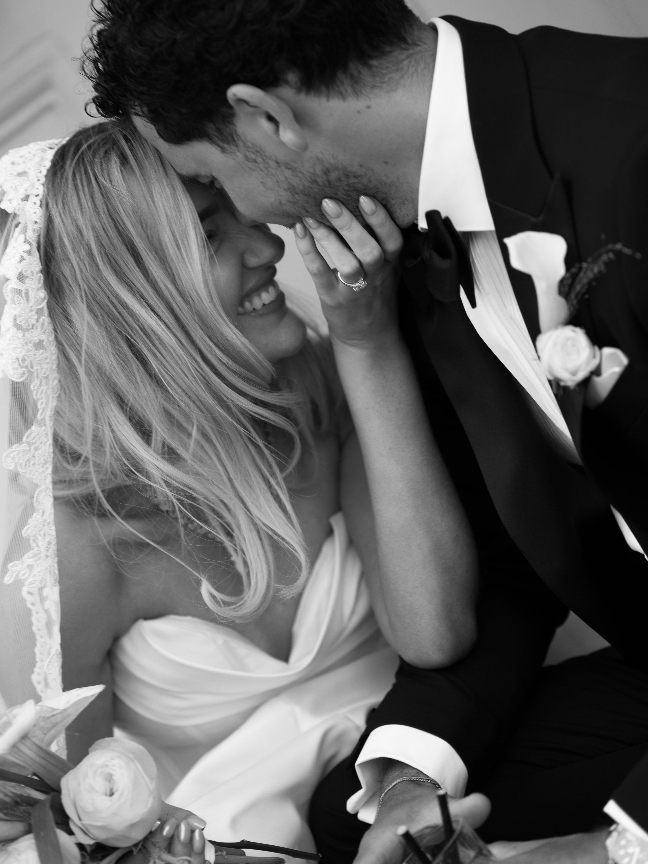 A black and white photo of a couple, the man wearing a tuxedo and the woman in a wedding dress, sharing a kiss with their foreheads touching. The woman has long, light-colored hair and the man has dark, curly hair. Their hands are visible as they hol