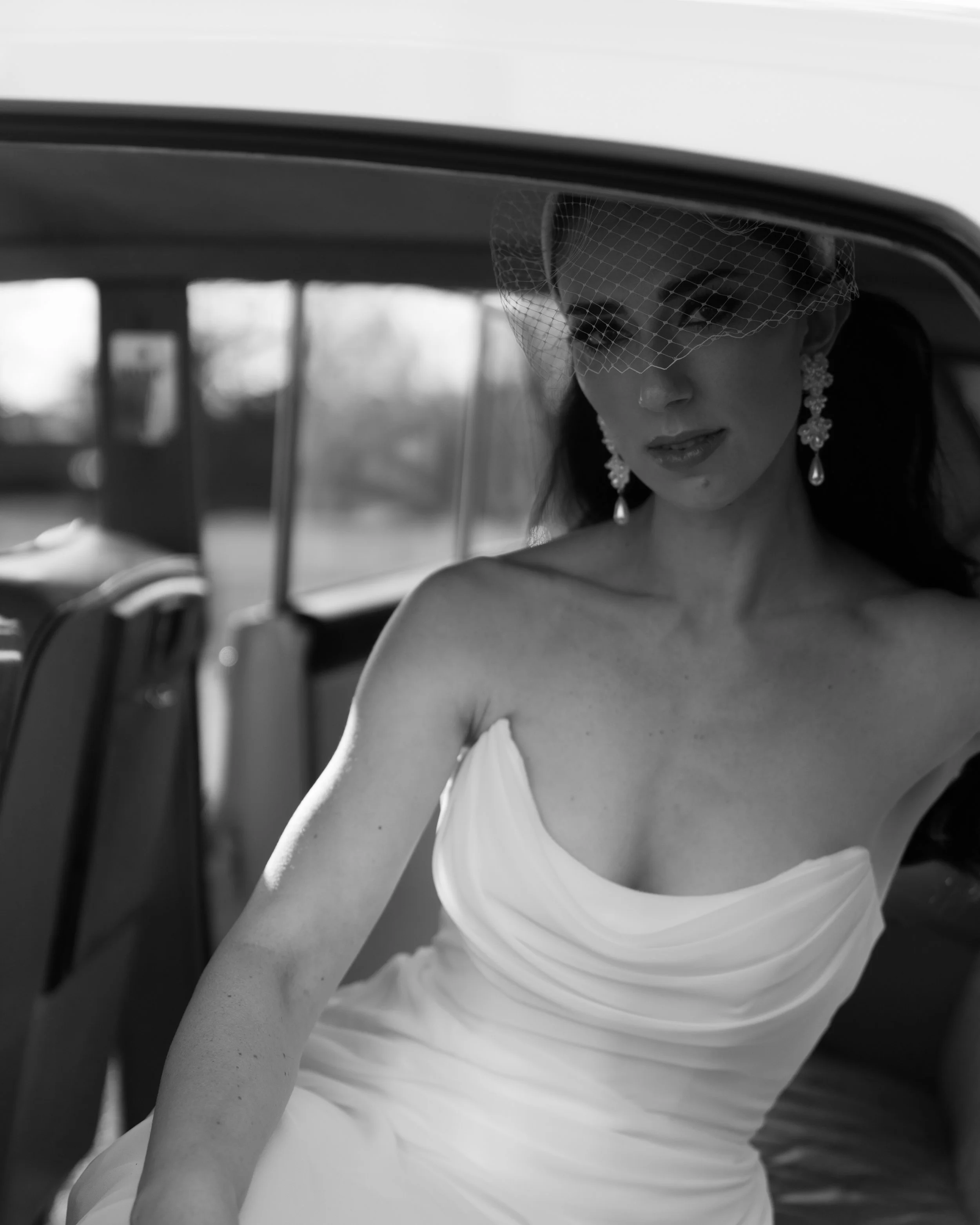 A black and white photo of a woman in a wedding dress sitting in a car. She is wearing earrings and a veil with a netted piece covering part of her face.