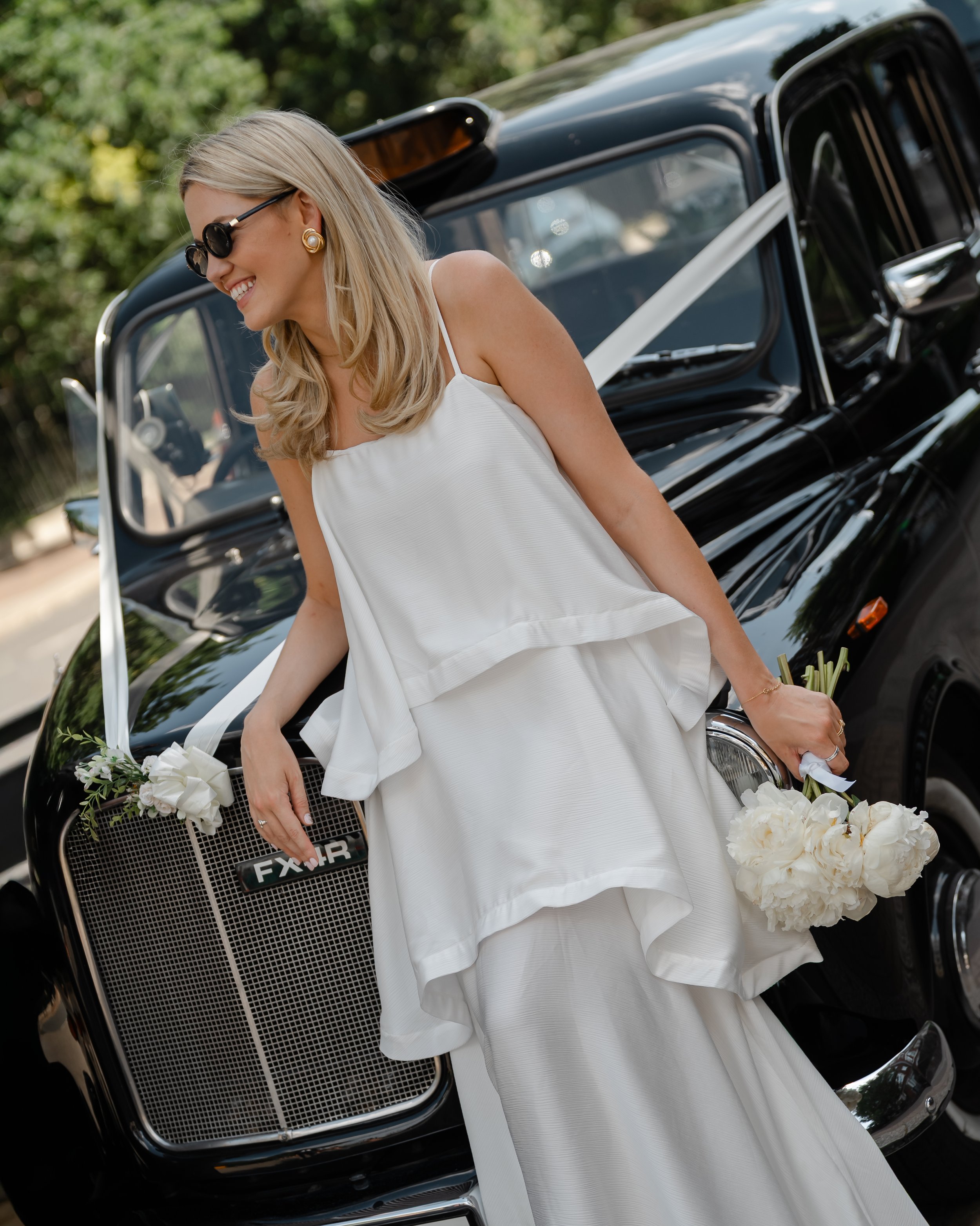Smiling woman in white dress holding a bouquet of white flowers in front of a vintage black car, wearing sunglasses and gold earrings.