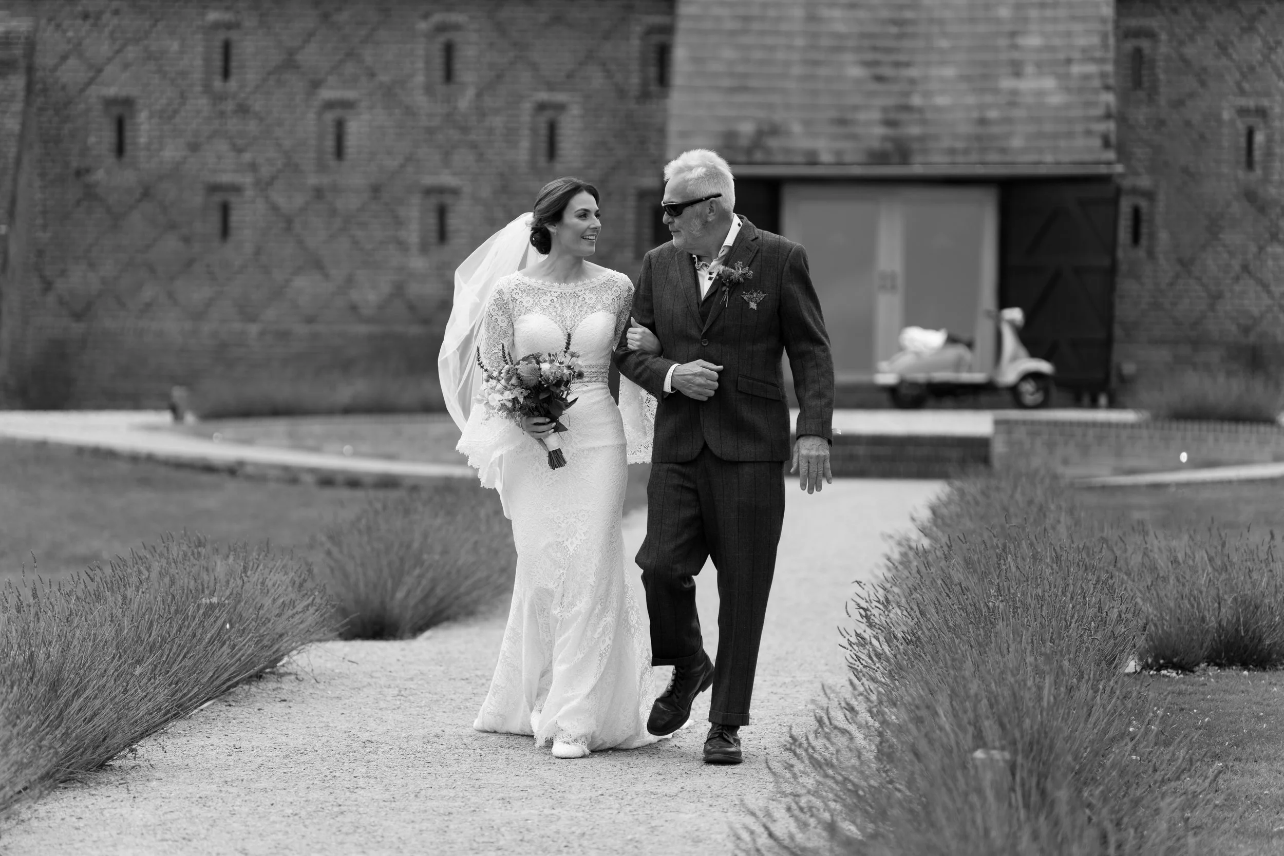 A bride in a lace wedding dress and veil walking arm-in-arm with a man in a dark suit, sunglasses, and a boutonniere, on a garden pathway near a brick building and a vintage car in the background.