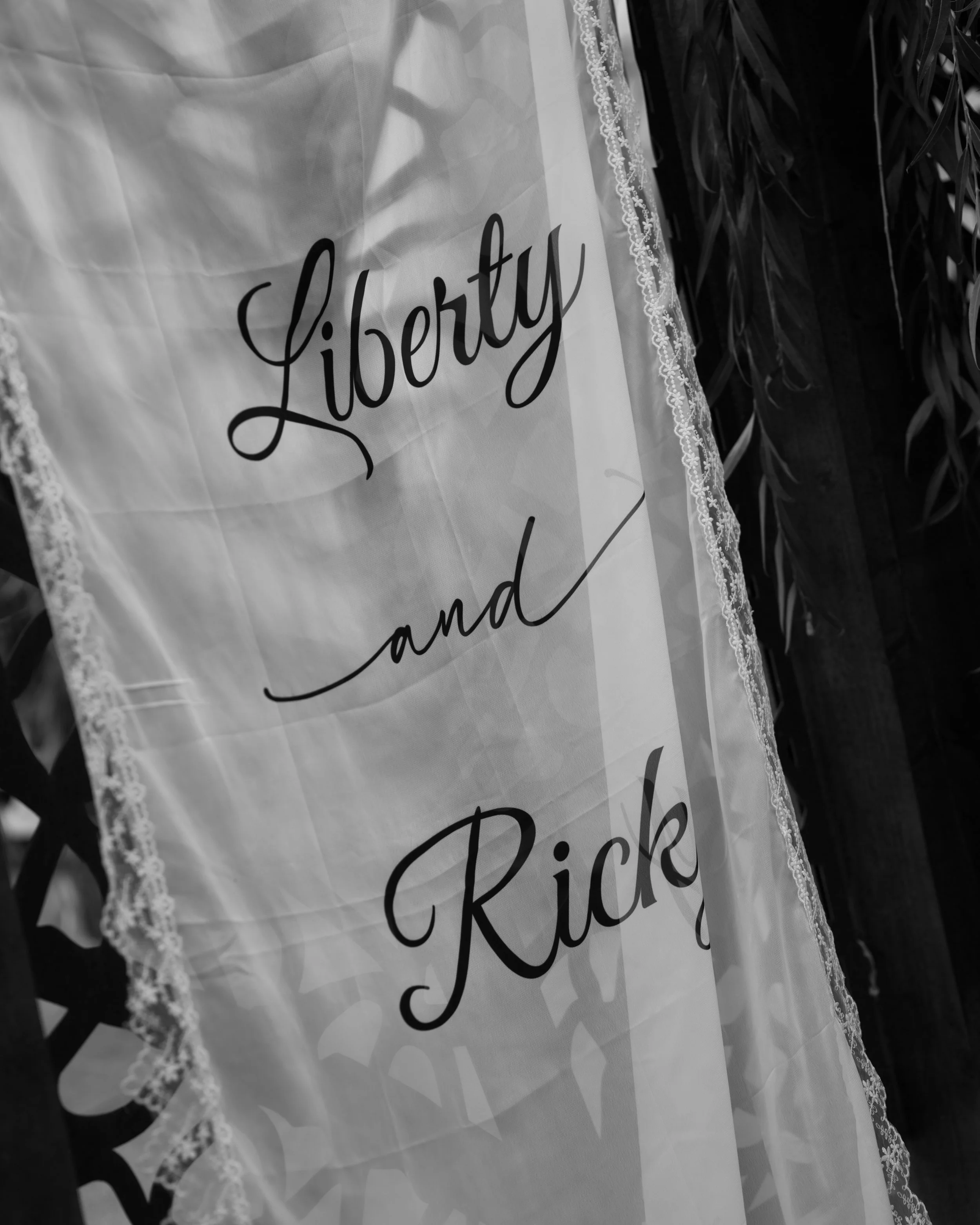 Close-up of a white sheer curtain with the words "Liberty and Pick" written on it in black cursive font, with lace trim along the edge. In the background, there are dark leaves or foliage.