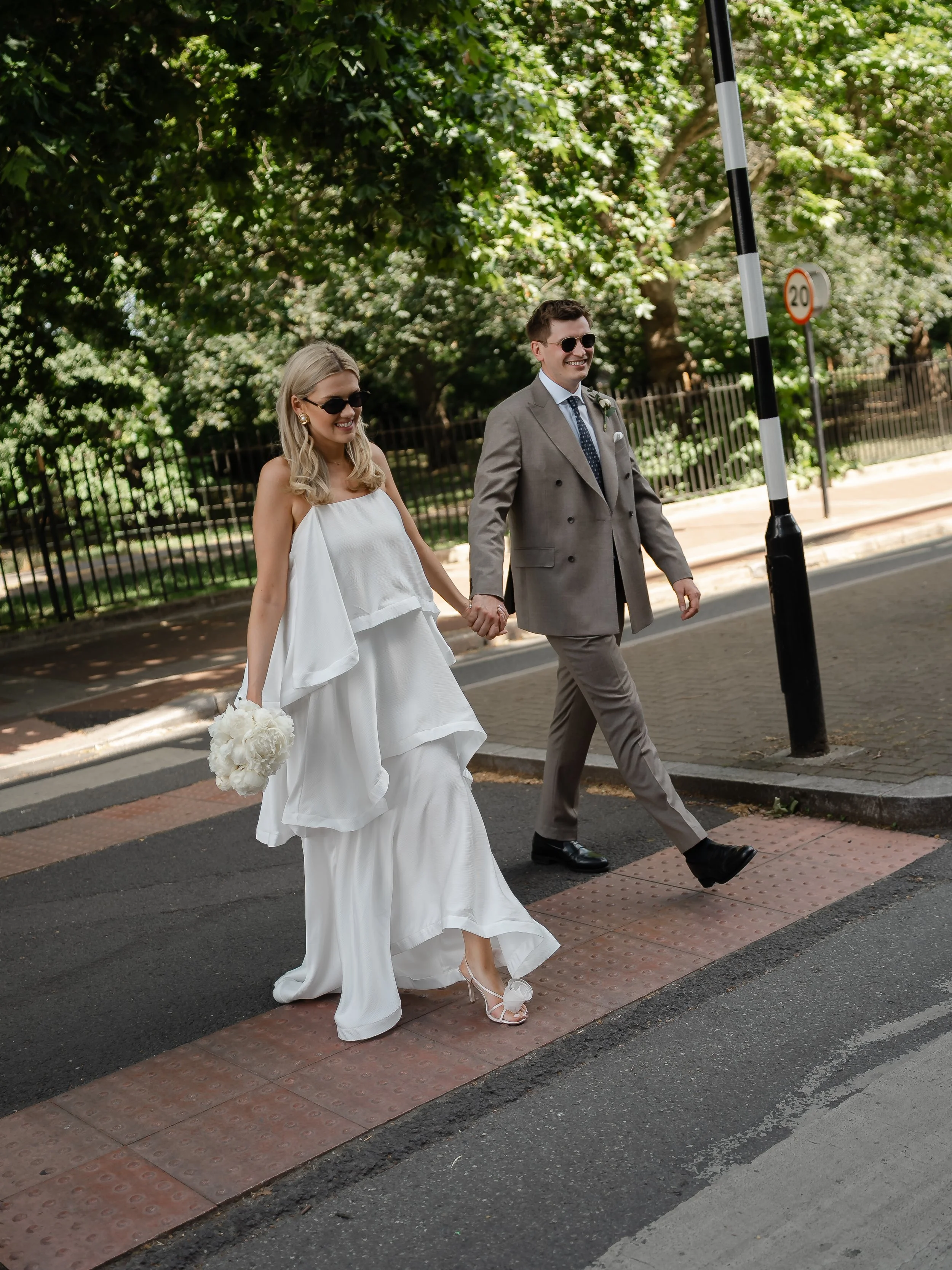 A newlywed couple walking hand in hand on a city street, smiling, with the woman in a white wedding dress holding a bouquet of white flowers, and the man in a gray suit and tie, both wearing sunglasses, with green trees and a black metal fence in the
