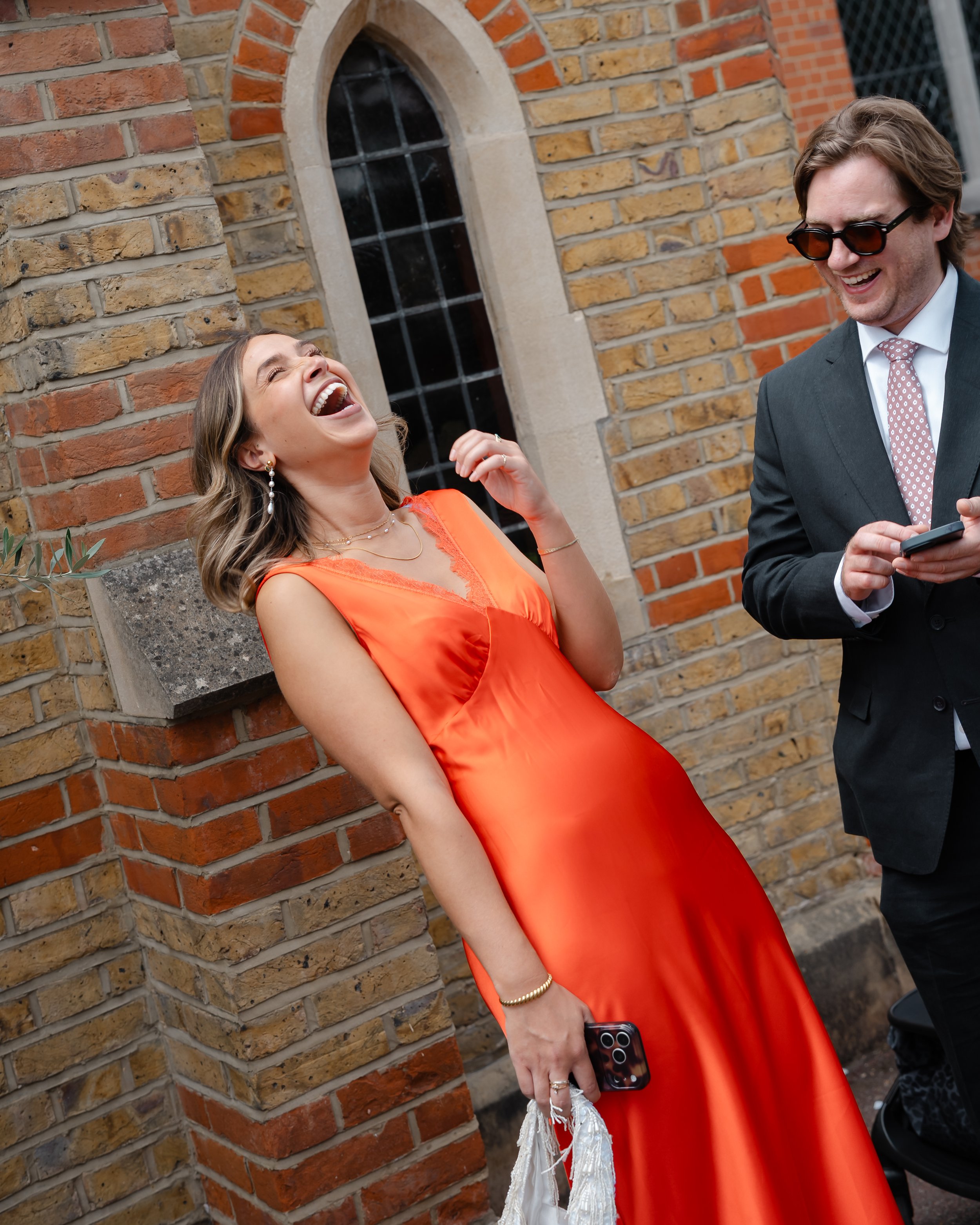 A woman in an orange dress is laughing while leaning against a brick wall, holding a phone and a white decorative item in her hand. A man in a dark suit with sunglasses is standing next to her, also smiling and looking at her.