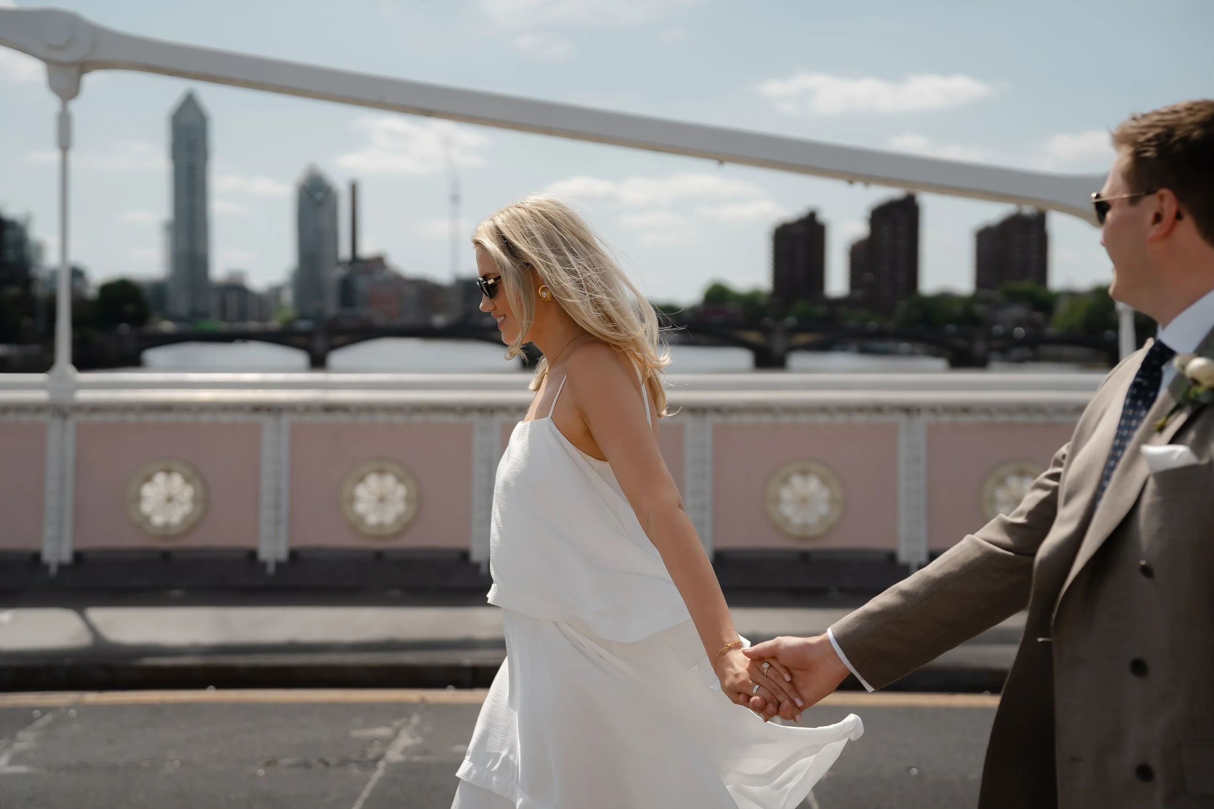 A bride and groom holding hands and walking outdoors on a bridge with a city skyline in the background, smiling.