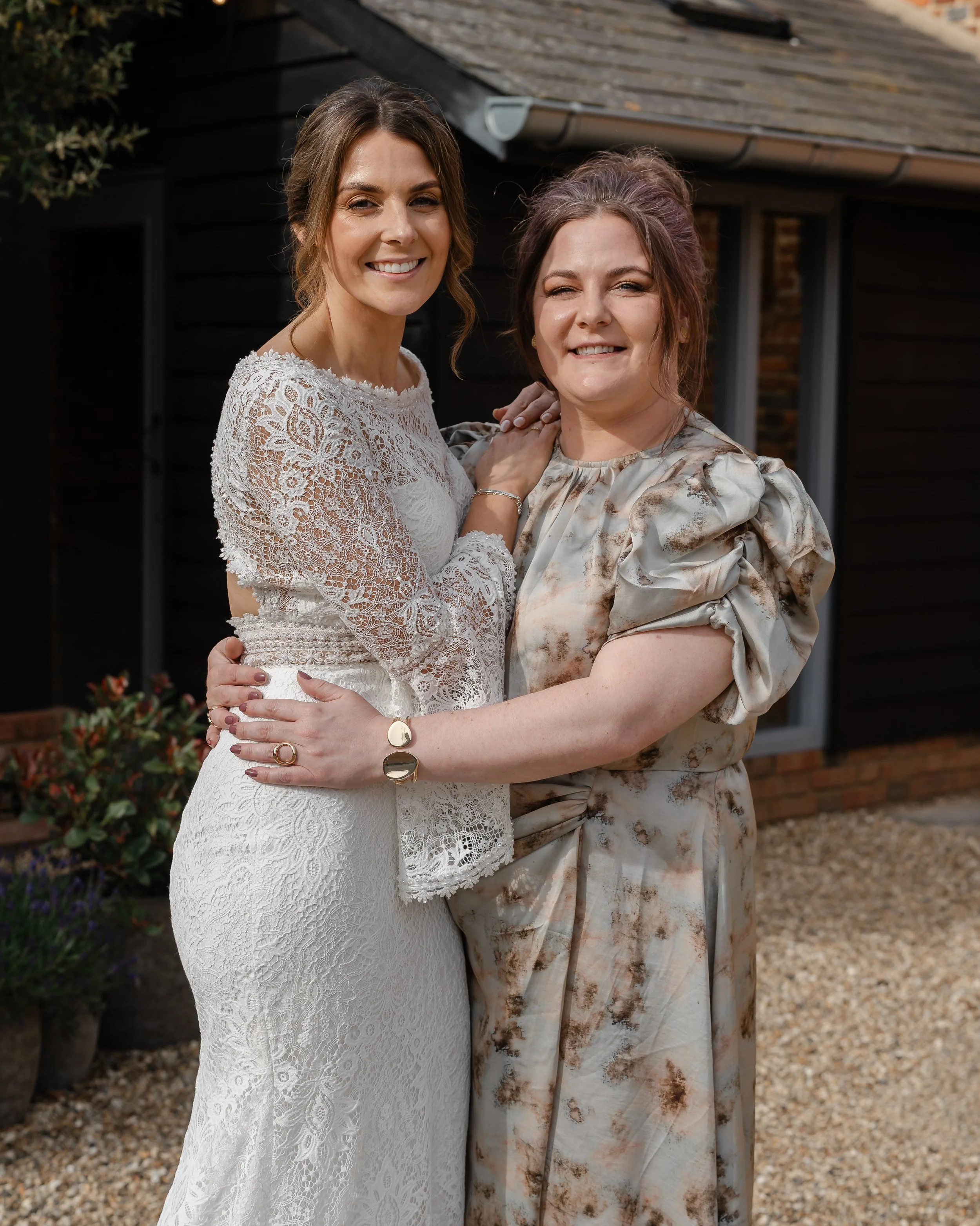 Two women hugging outdoors in front of a house, one in a white lace dress and the other in a patterned dress with puffy sleeves, both smiling.