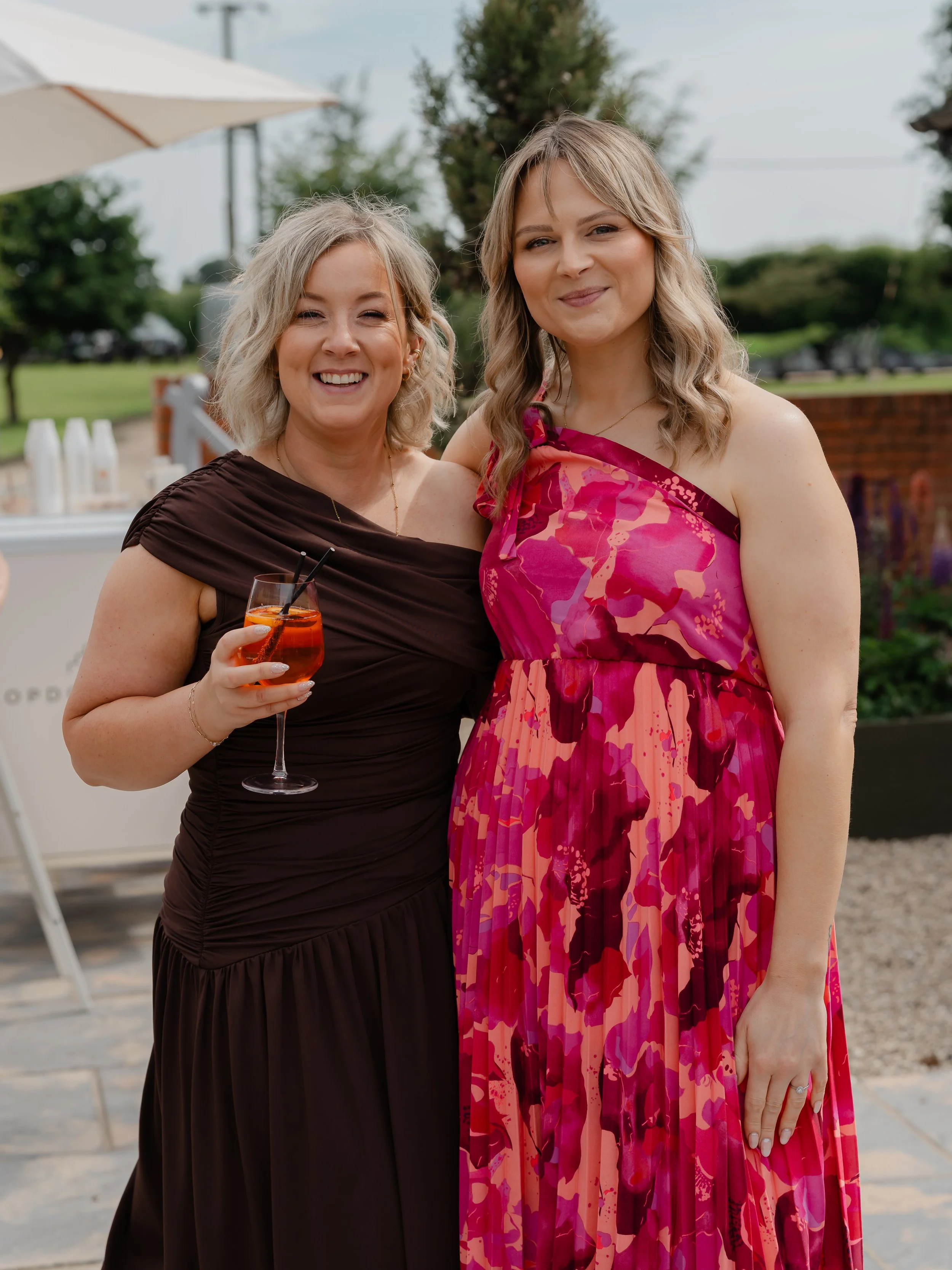 Two women standing outdoors, smiling at the camera, one holding a drink with an orange liquid and ice. The woman on the left wears a dark brown dress with one shoulder strap, and the woman on the right wears a colorful pink and purple floral dress. T