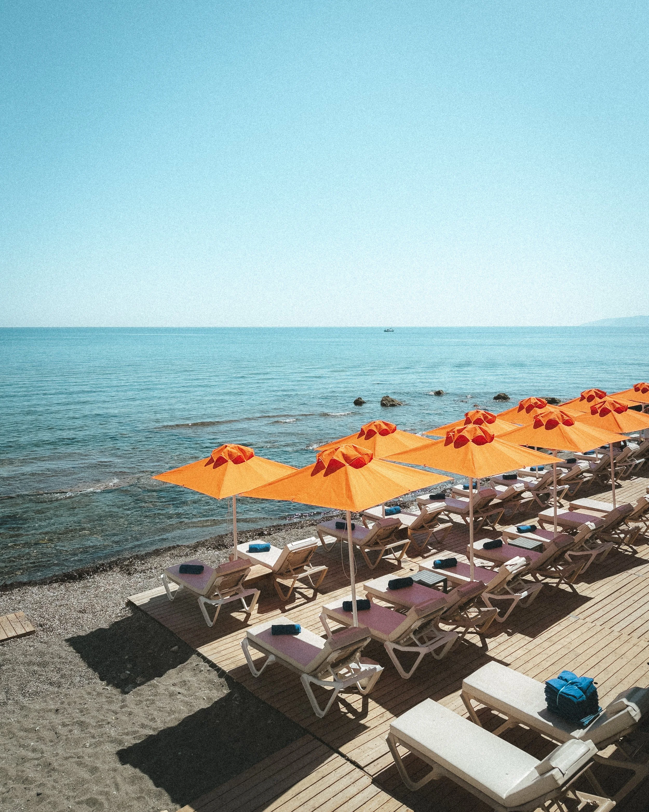 Row of white lounge chairs with blue towels and yellow umbrellas on a wooden deck by the sea, with a boat in the distance.