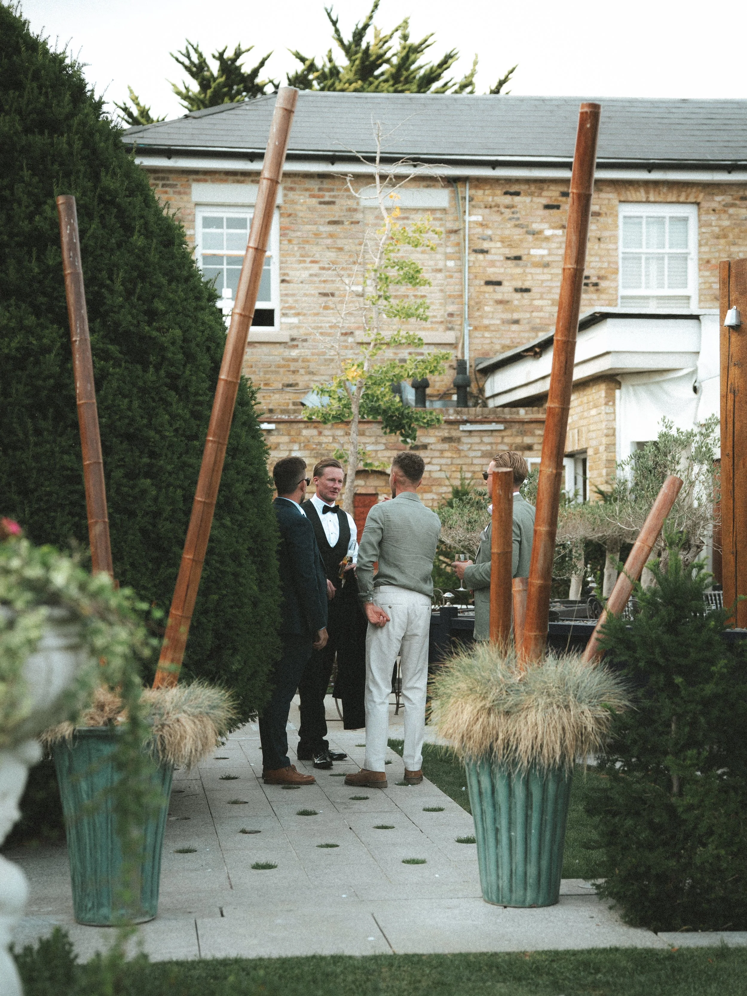 People in formal attire talking at an outdoor event in a garden with large potted plants, a brick house in the background, and tall wooden poles.