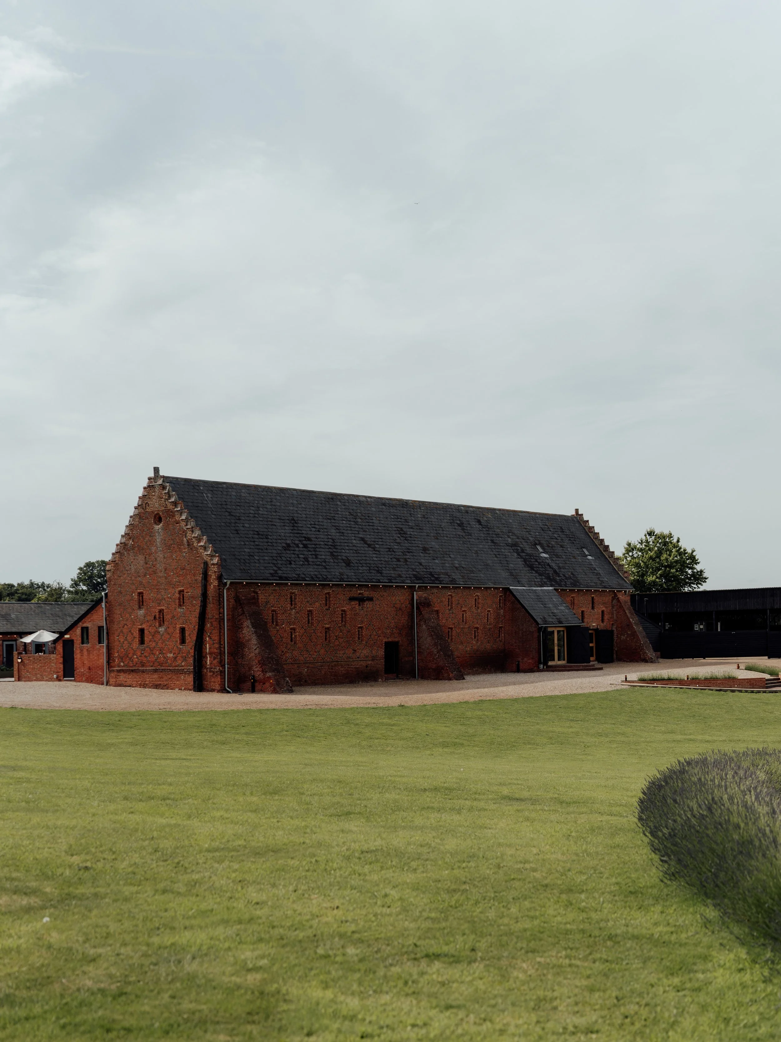 A historic red brick barn with a sloped roof on a green lawn, under a cloudy sky at Copdock Hall, Suffolk.