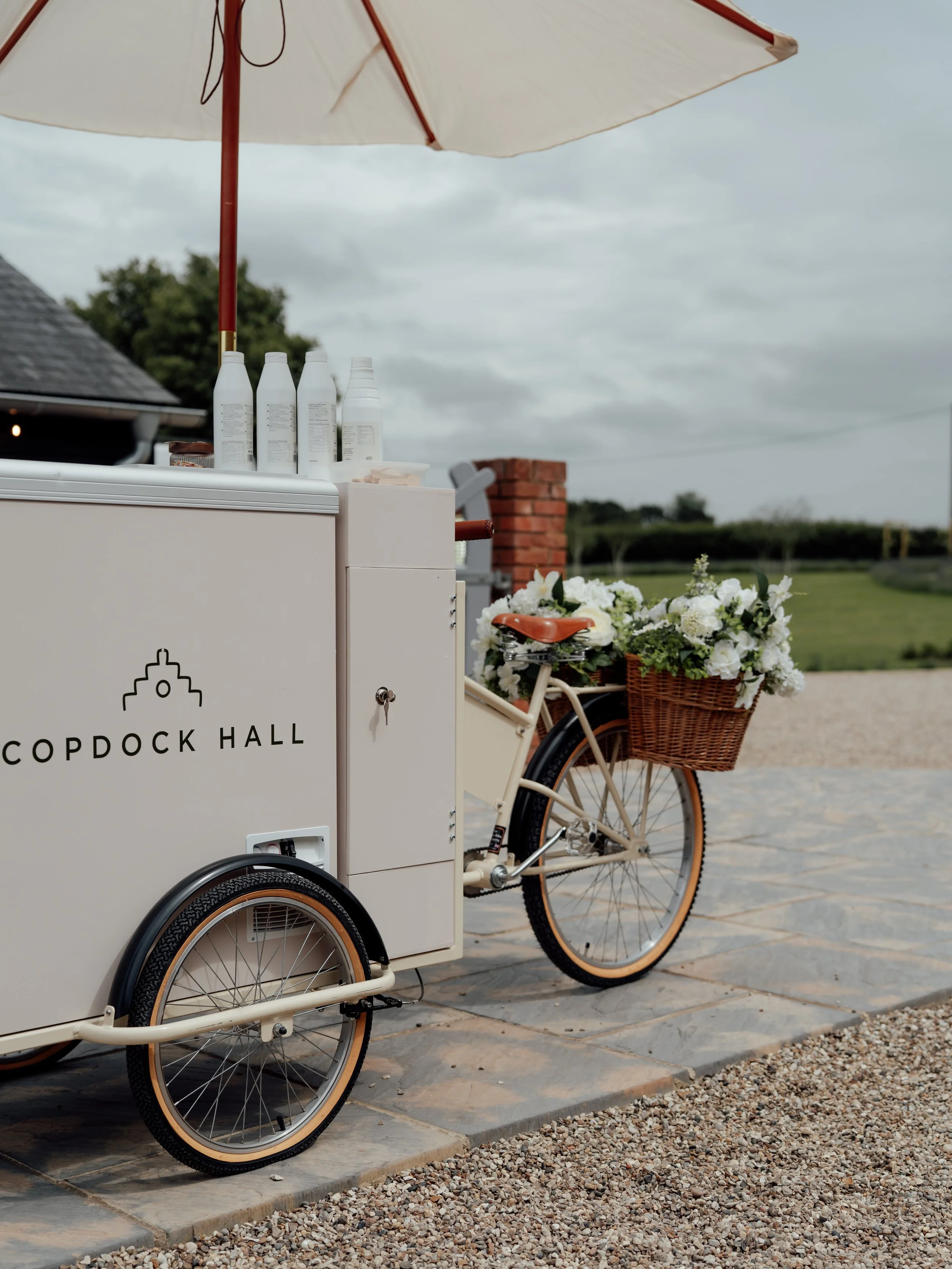 A white bicycle with a brown wicker basket filled with white flowers, parked next to a white beverage cart labeled 'Copdock Hall,' underneath a large beige patio umbrella on a cloudy day.