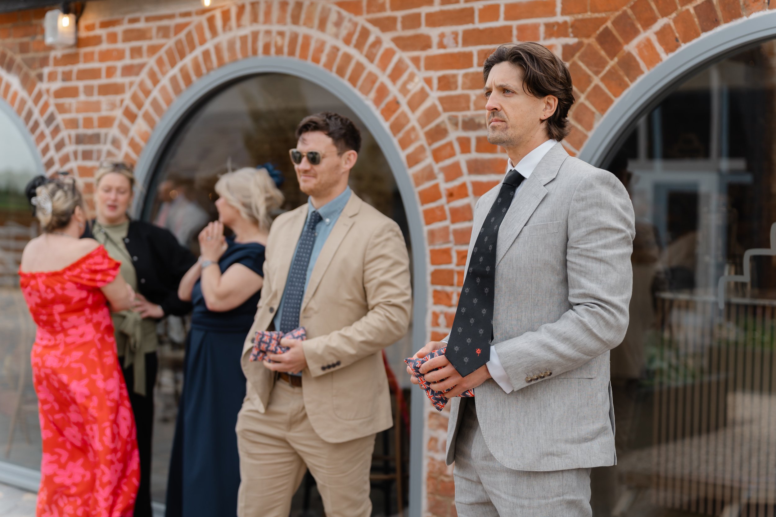 A group of people dressed in formal attire gathering outside a brick building, with some engaged in conversation and one man standing apart looking serious.