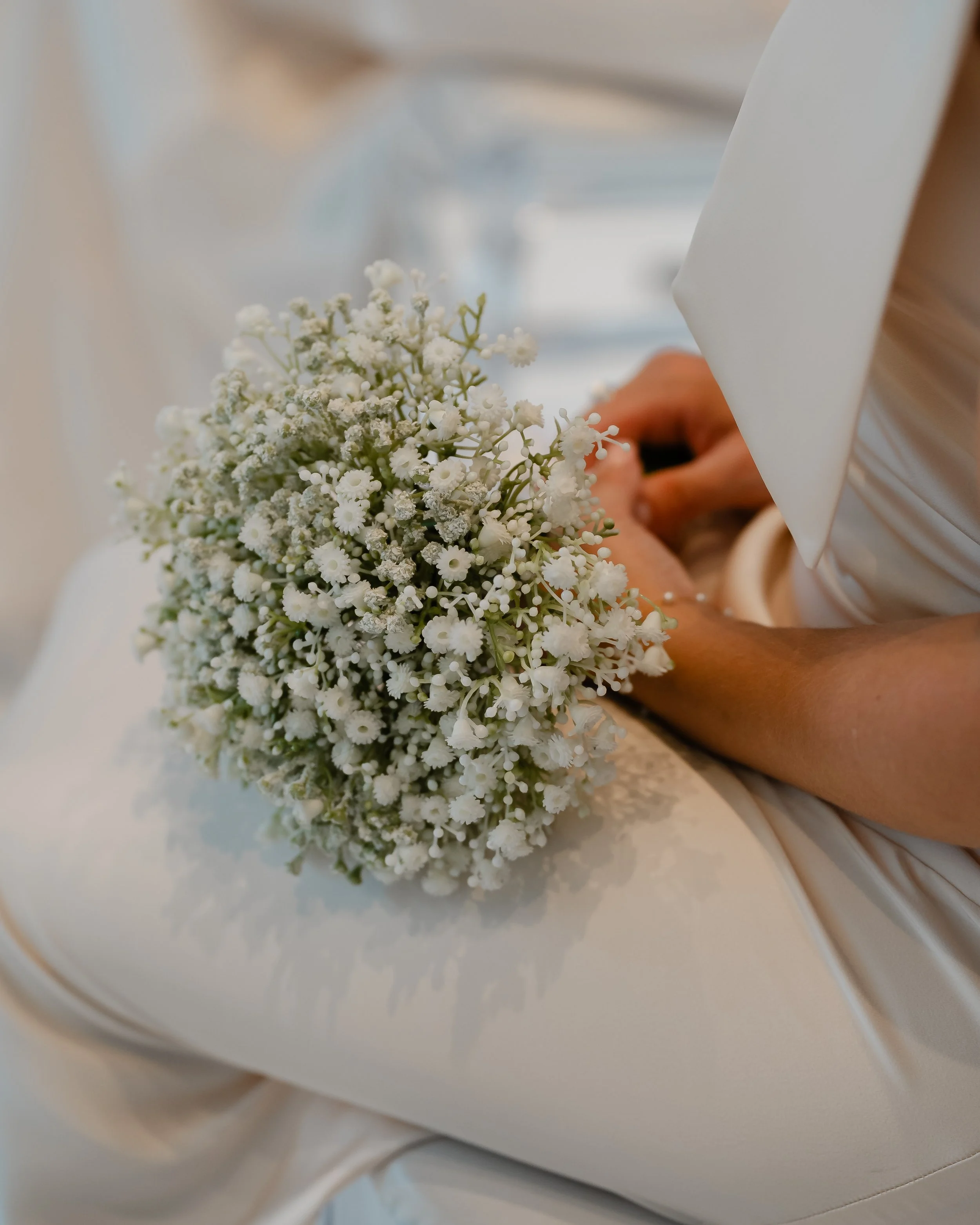 A person is holding a small bouquet of white flowers with delicate, tiny blooms in white and green. The person’s hand is visible, and they are wearing a white long-sleeved garment. The background is blurred, focusing on the flowers and the hand.