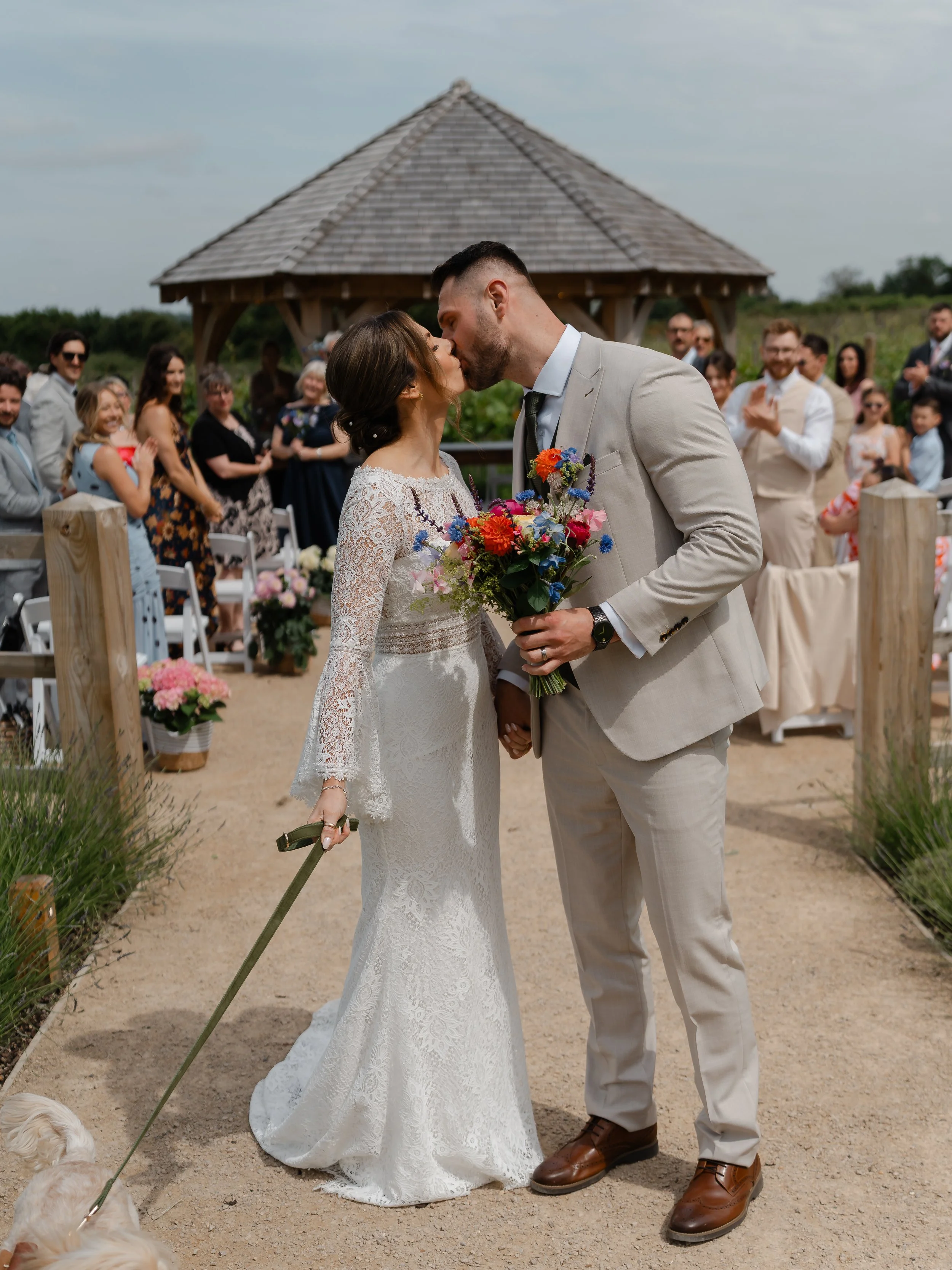A bride and groom kiss during their outdoor wedding ceremony, surrounded by guests, with a gazebo in the background.