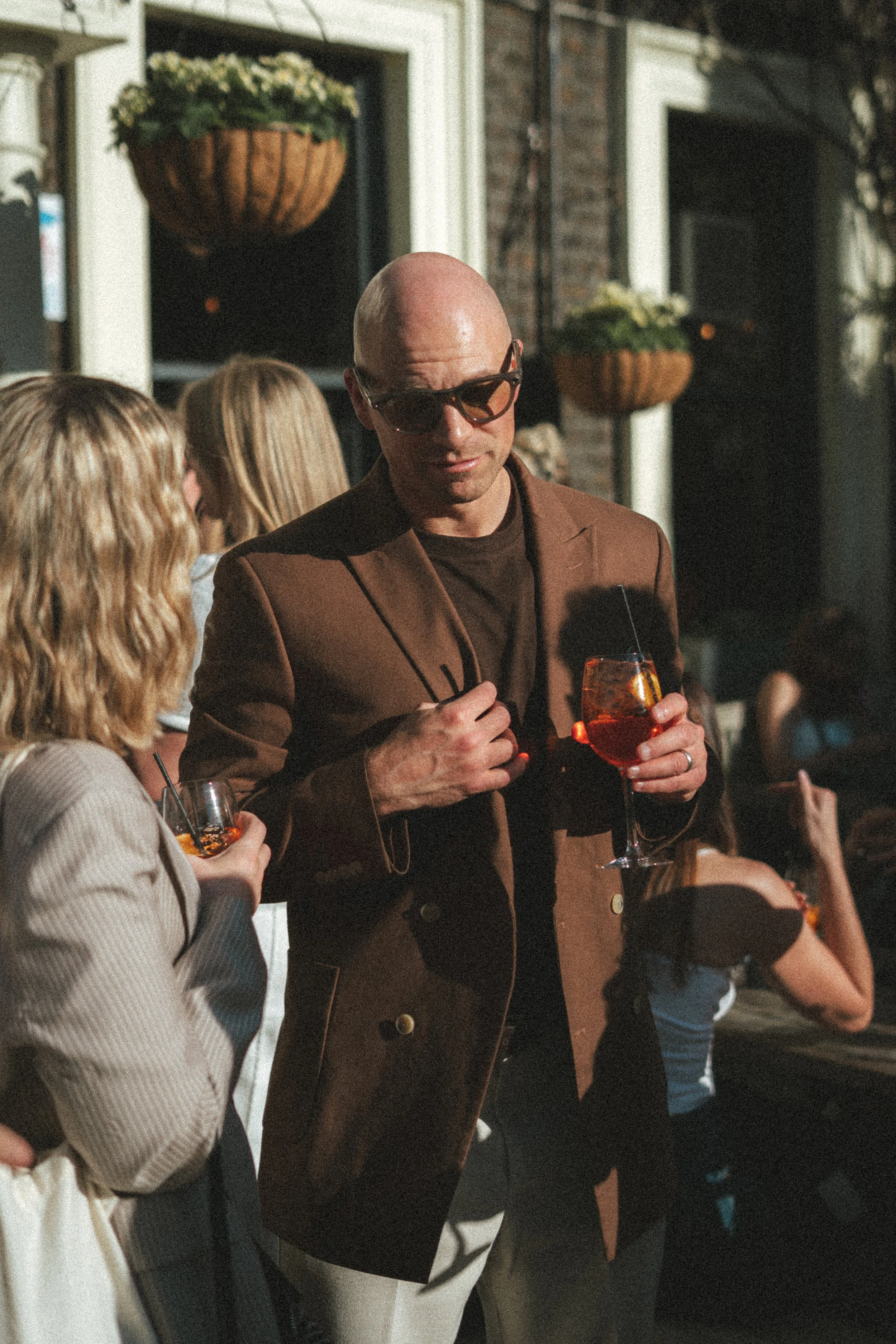 A man wearing sunglasses and a brown blazer holding a glass of an orange-colored drink at an outdoor gathering in the evening, with other people and hanging flower baskets in the background.