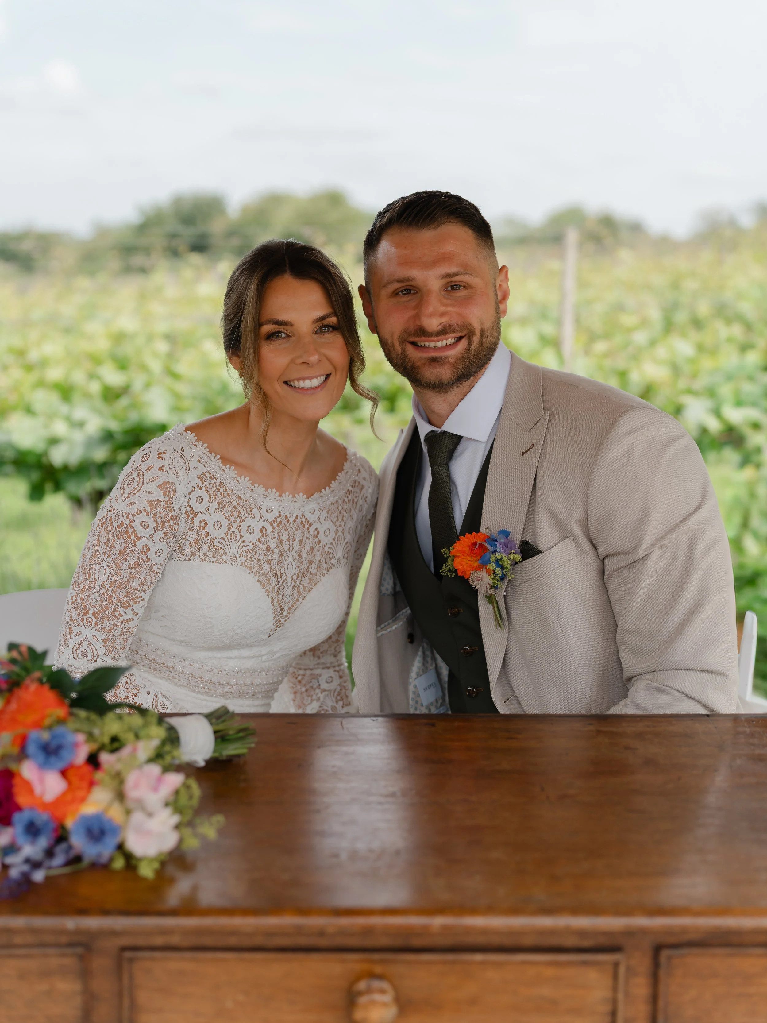 A smiling woman in a white lace wedding dress standing next to a smiling man in a light-colored suit and black tie, both posing outdoors with a blurred green landscape background.