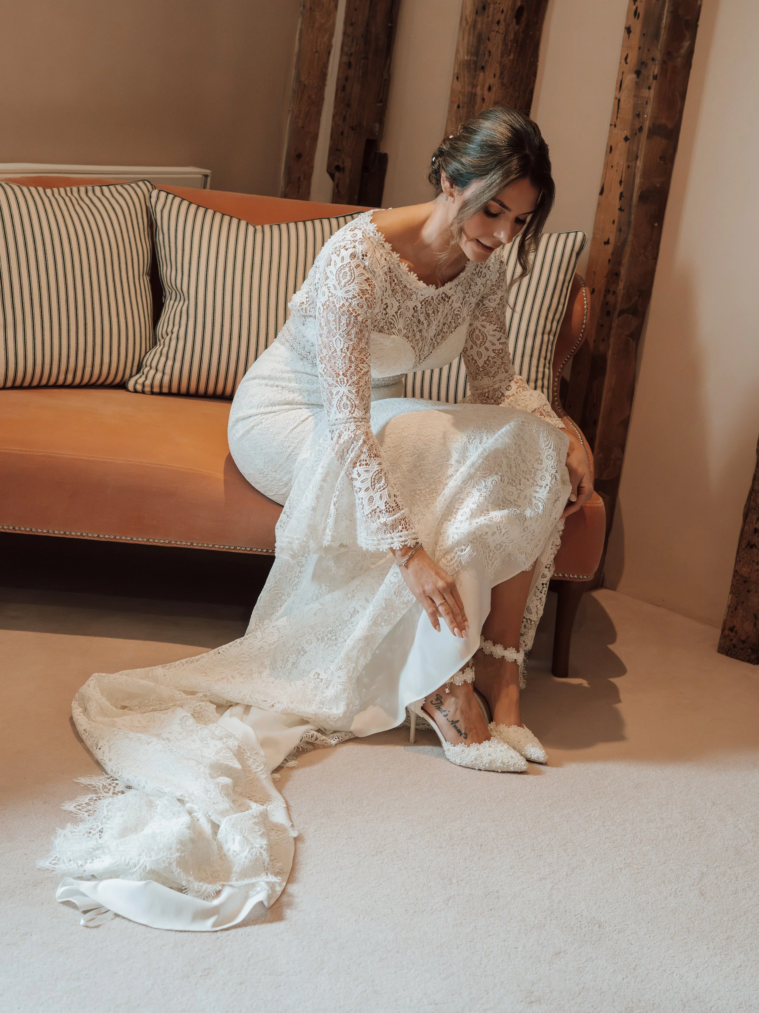 A woman dressed in a white lace wedding dress, sitting on a striped sofa, adjusting her white lace high heels, with her hair styled in loose waves.