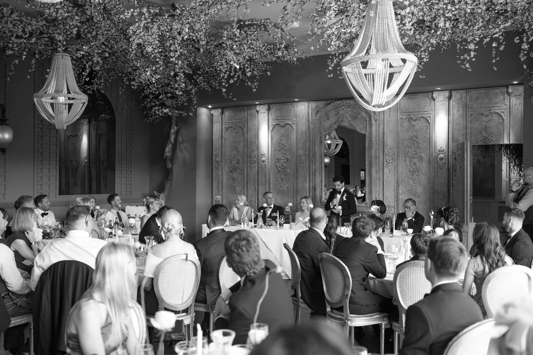 Black and white photo of a formal dinner event with many people seated at tables, a man in a tuxedo giving a speech, ornate walls, large chandeliers, and floral decorations overhead.