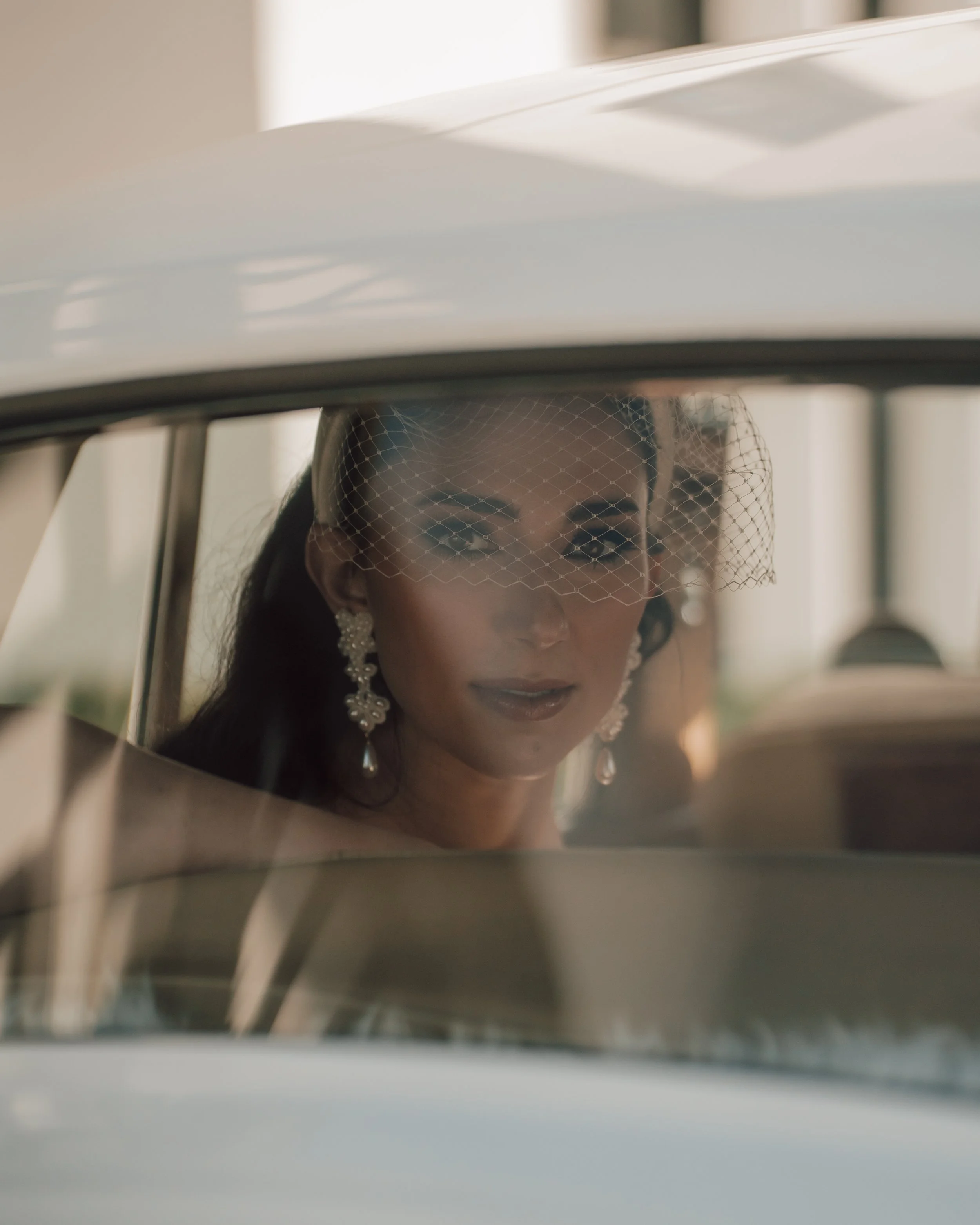 Woman with dark hair, wearing pearl earrings and a birdcage veil, looking through a car window.
