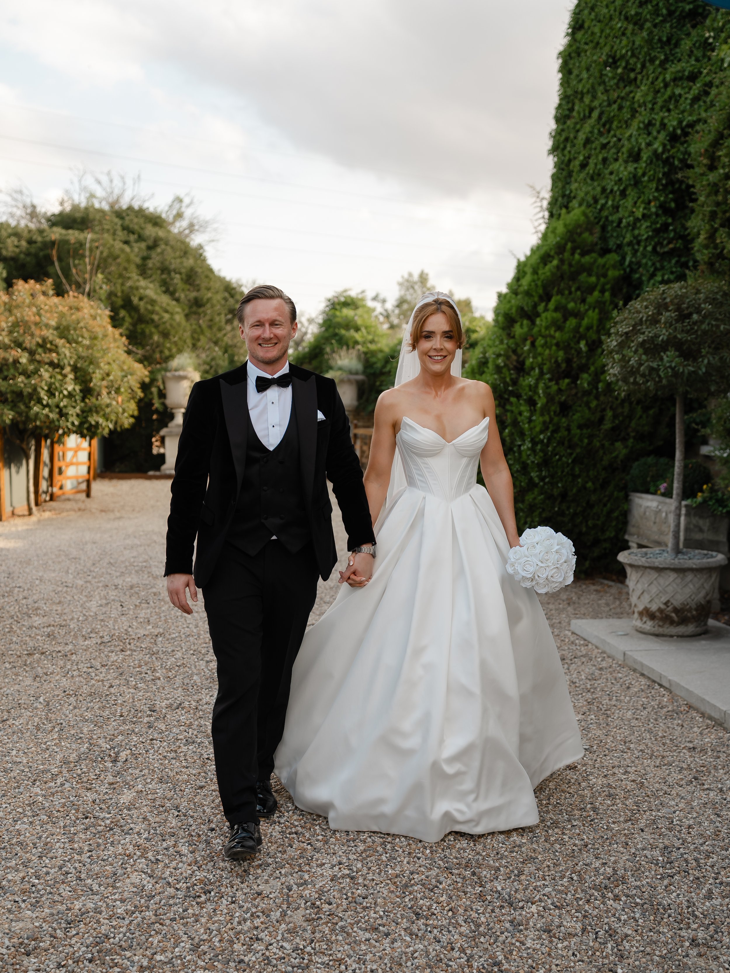 A newlywed couple walking outdoors on a gravel path, holding hands and smiling. The groom is dressed in a black tuxedo with a bow tie, and the bride is wearing a strapless white wedding gown with a full skirt and holding a bouquet of white roses.