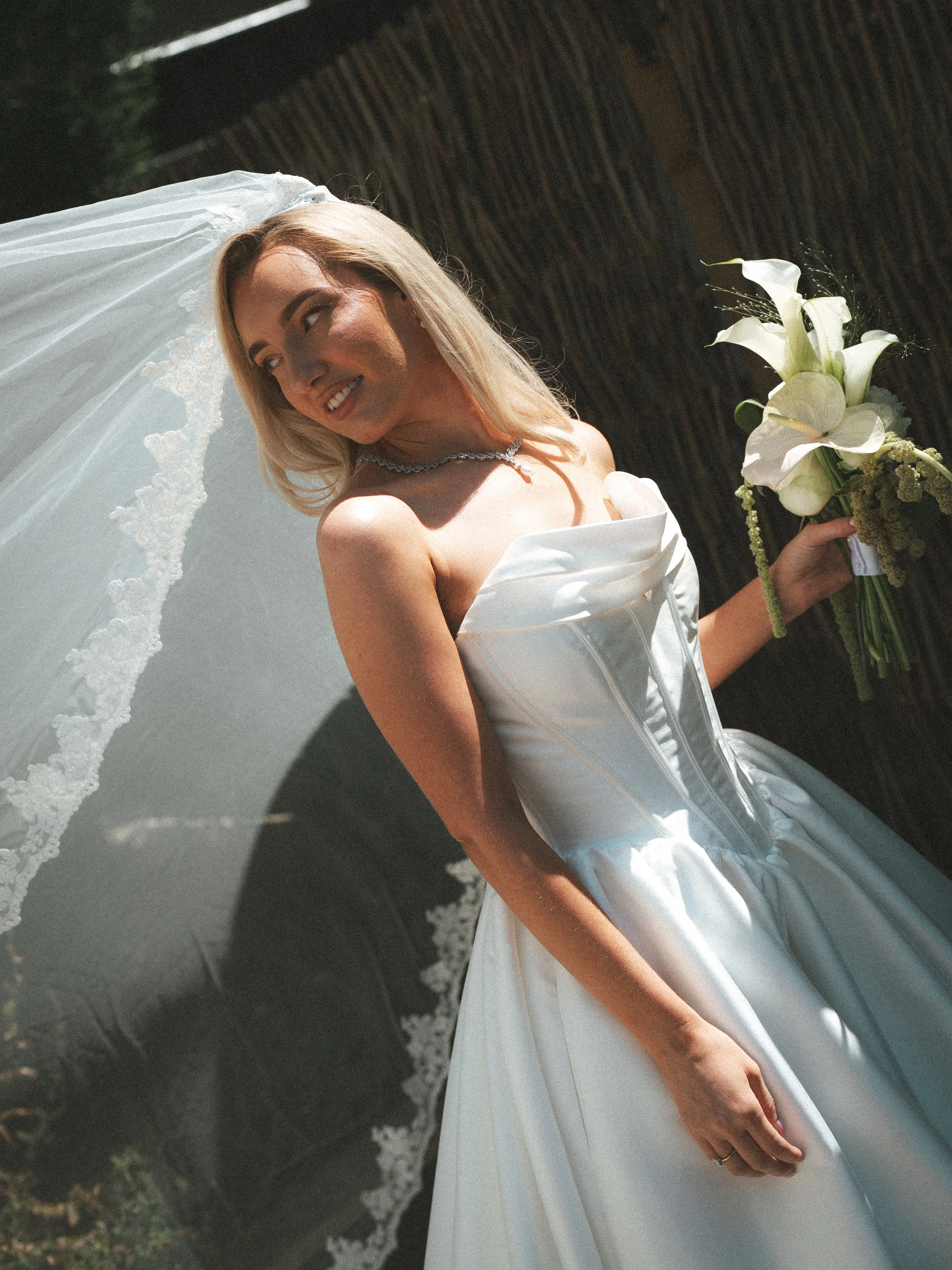 A bride with blonde hair, wearing a white strapless wedding dress and a veil, holding a bouquet of white calla lilies, standing outdoors with greenery and a wooden fence in the background.