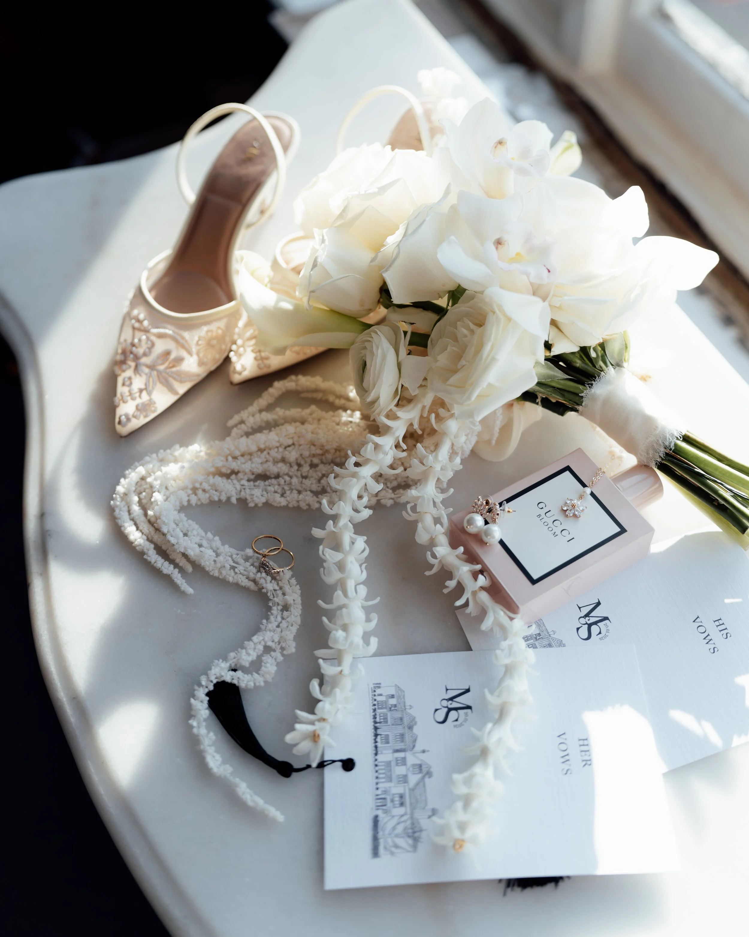 A wedding bouquet with white flowers, peeking out of a Gucci Bloom perfume box, gold jewelry including earrings and rings, a pearl necklace, ivory high-heeled shoes with floral embroidery, and various wedding stationery on a white surface near a wind
