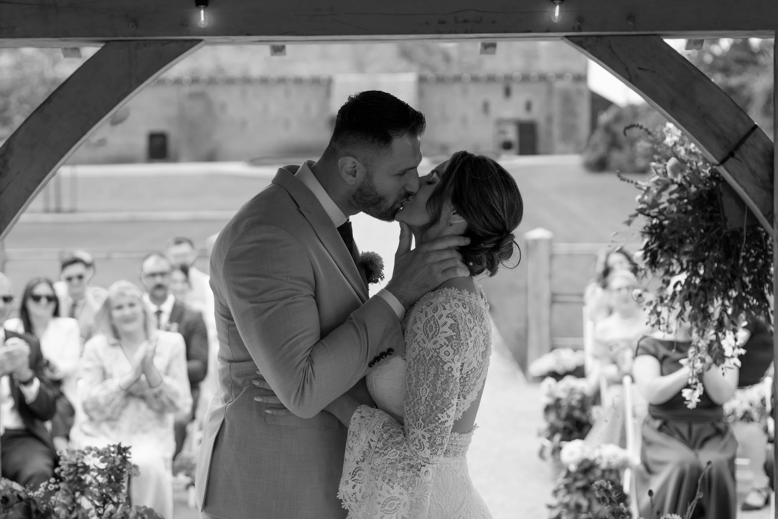 A bride and groom share a kiss during their wedding ceremony outdoors, framed by a wooden archway, with guests seated behind them.