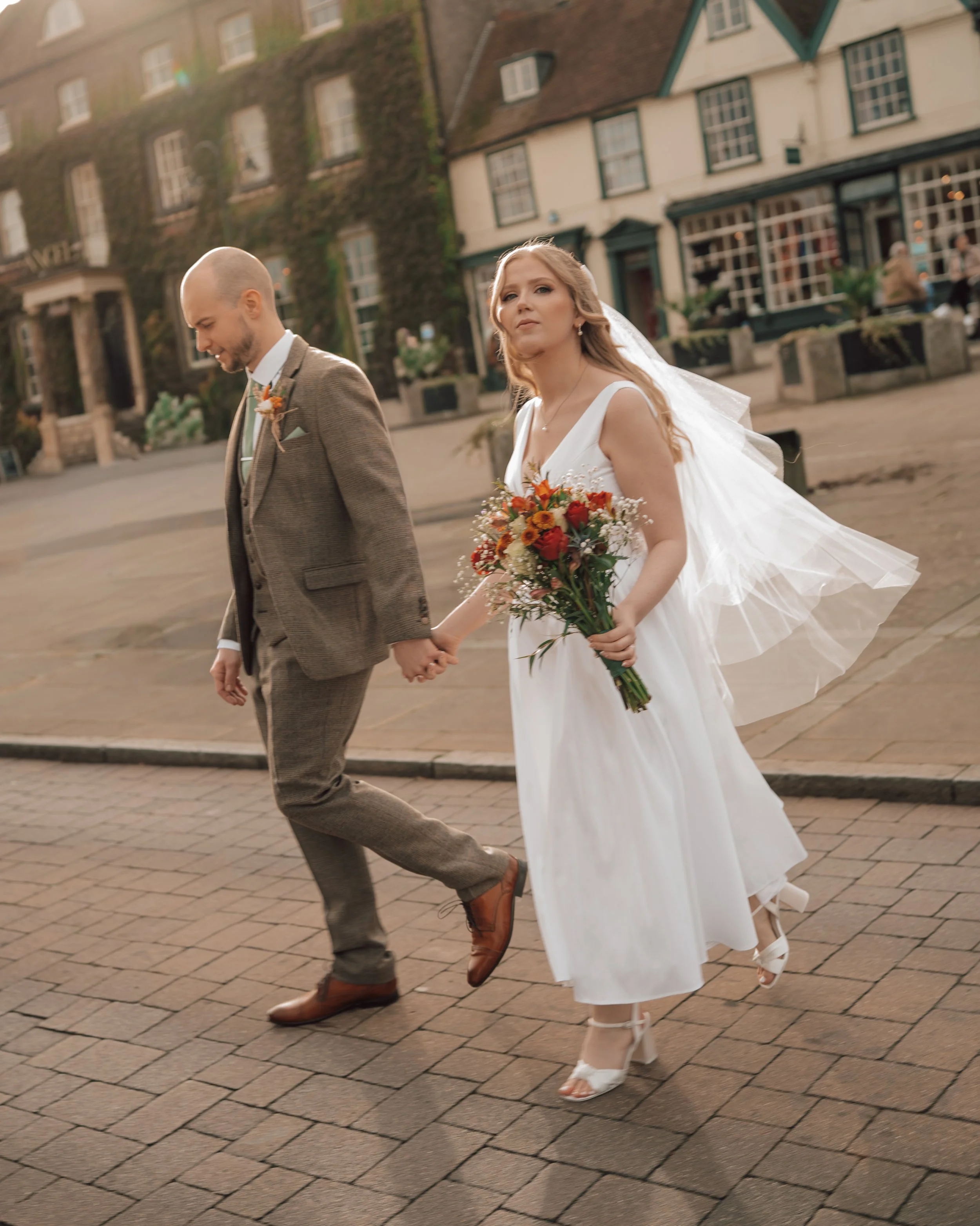 A bride and groom walking hand in hand on a city street, holding a bouquet, with historic buildings in the background.