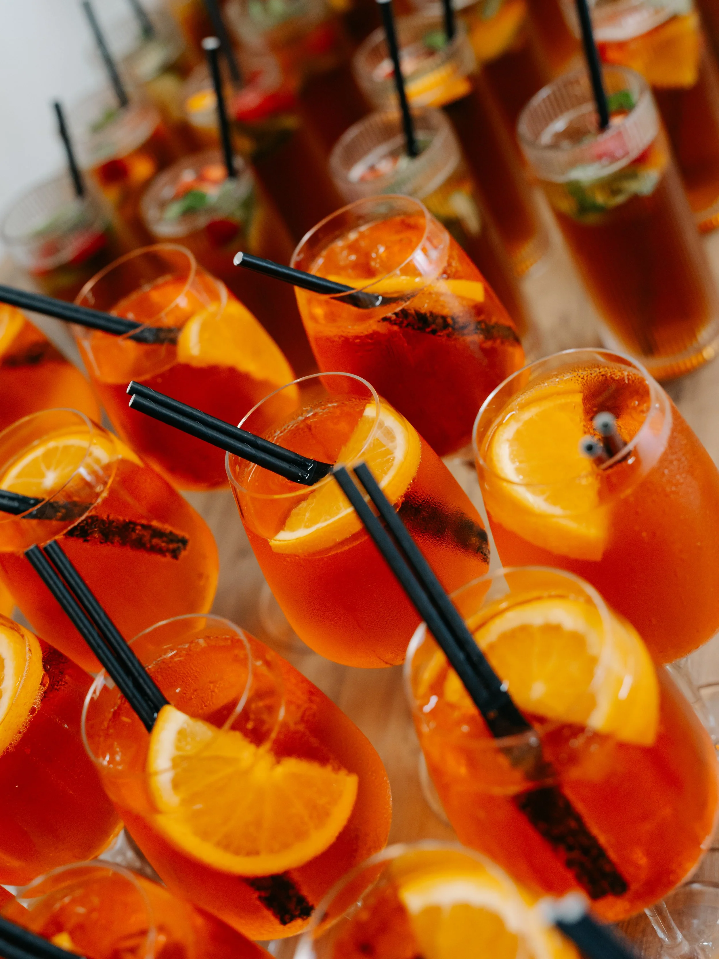 Close-up of multiple glasses of orange-colored drinks garnished with orange slices and mint, with black straws.