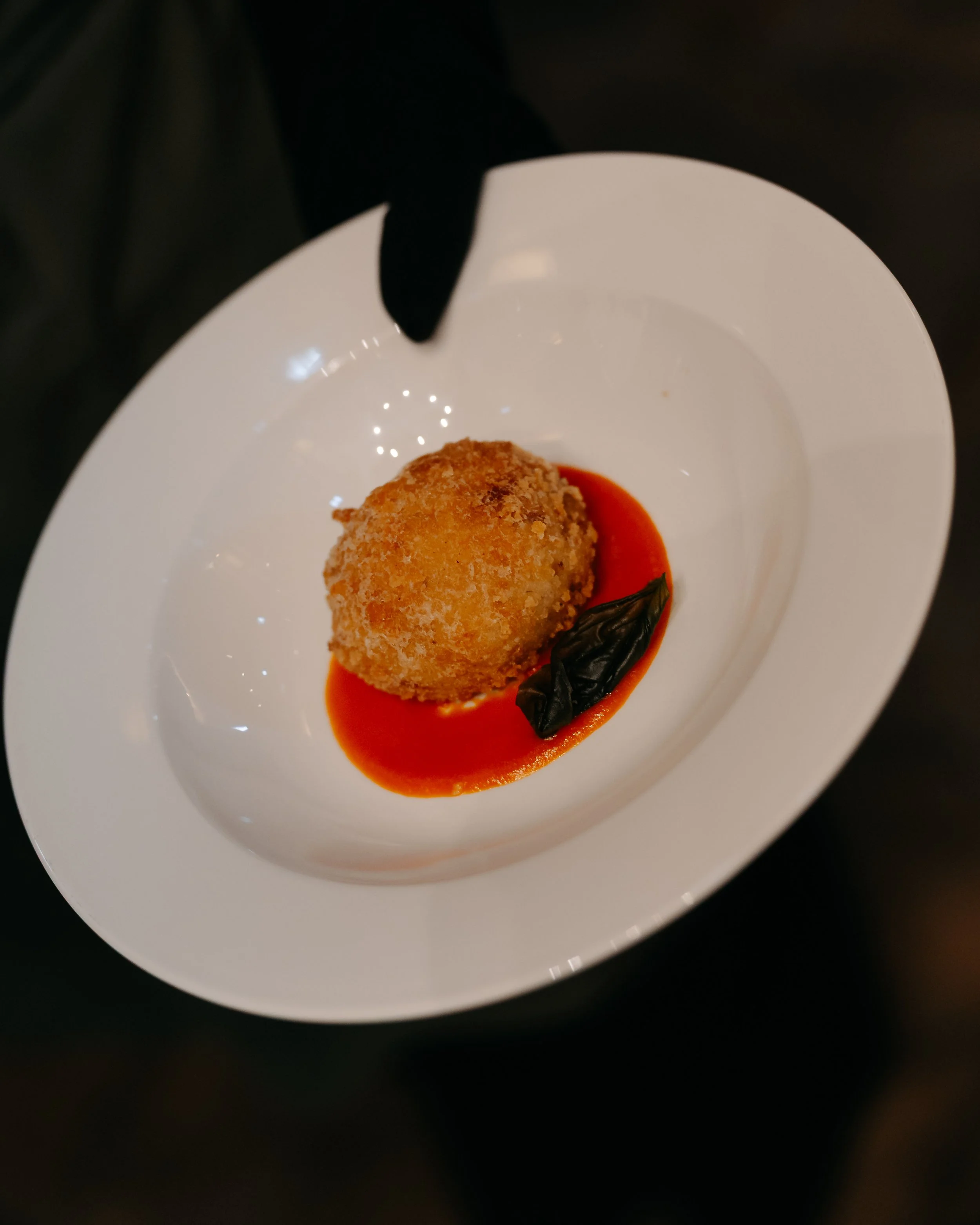 A breaded, fried appetizer or dessert on a white plate, garnished with a dark basil leaf and surrounded by red sauce.
