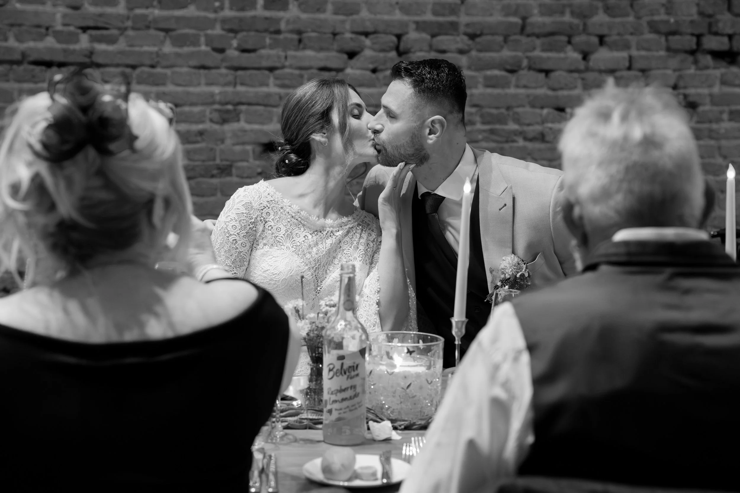 A bride and groom share a kiss at their wedding reception, sitting at a table with wedding decor, candles, and beverages, with a brick wall in the background.