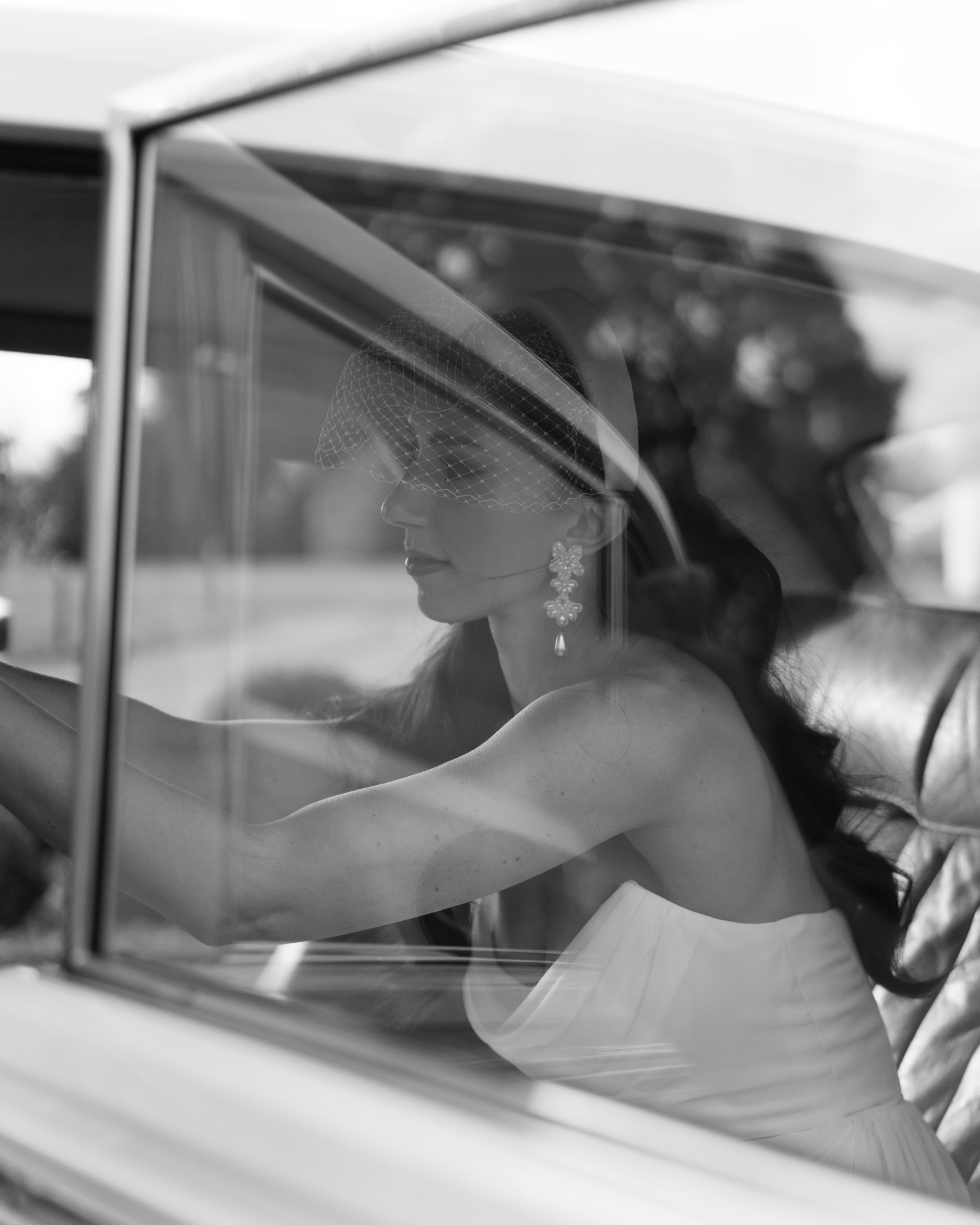 Black and white photo of a woman in vintage attire sitting inside a car, wearing a lace hat with a veil and large earrings, seen through the car window.