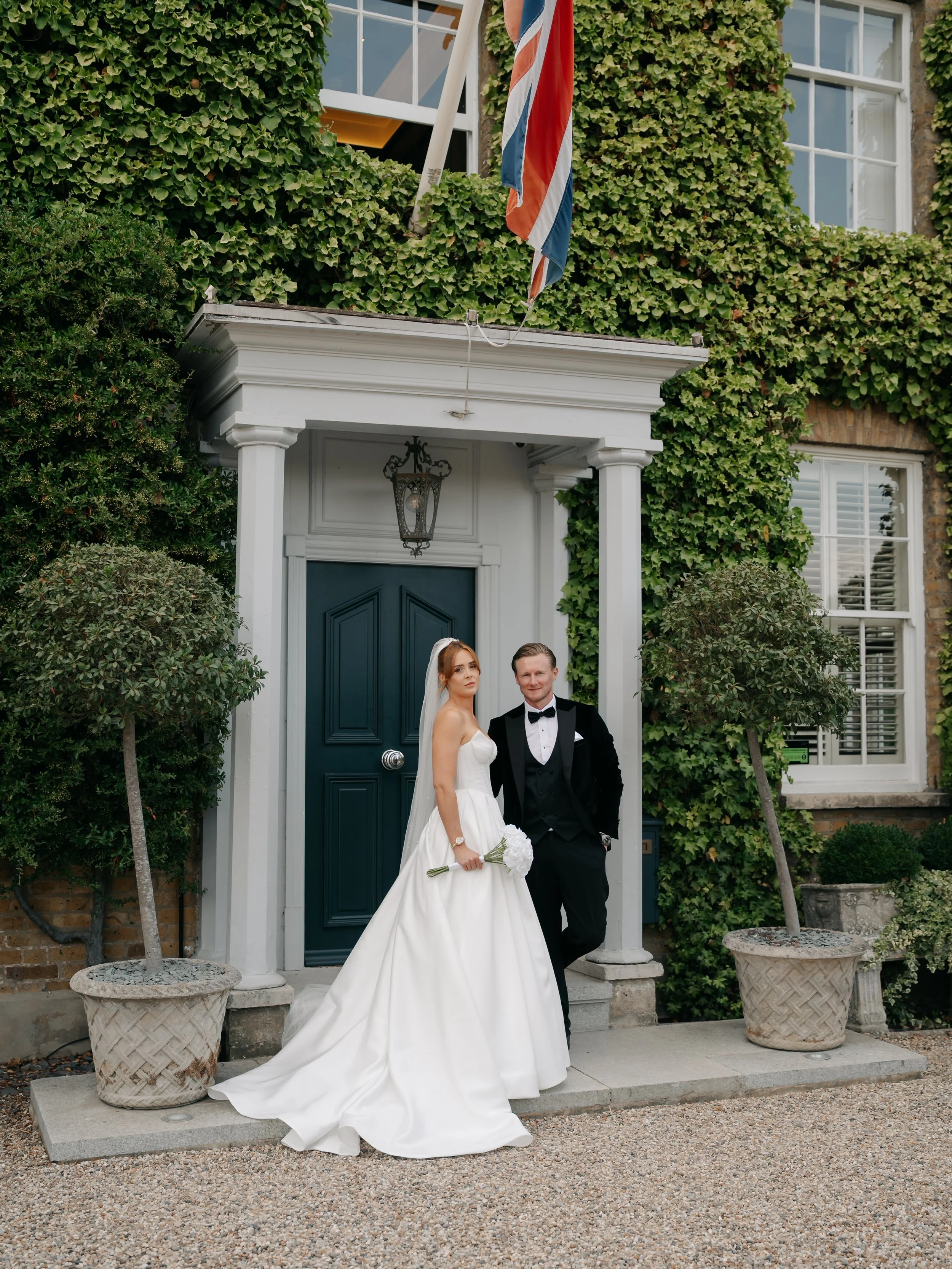 A bride and groom standing on steps in front of a house with ivy-covered walls, under a white porch roof with a hanging lantern. The bride is in a white wedding dress holding a bouquet, and the groom is in a tuxedo. A Union Jack flag is hanging above
