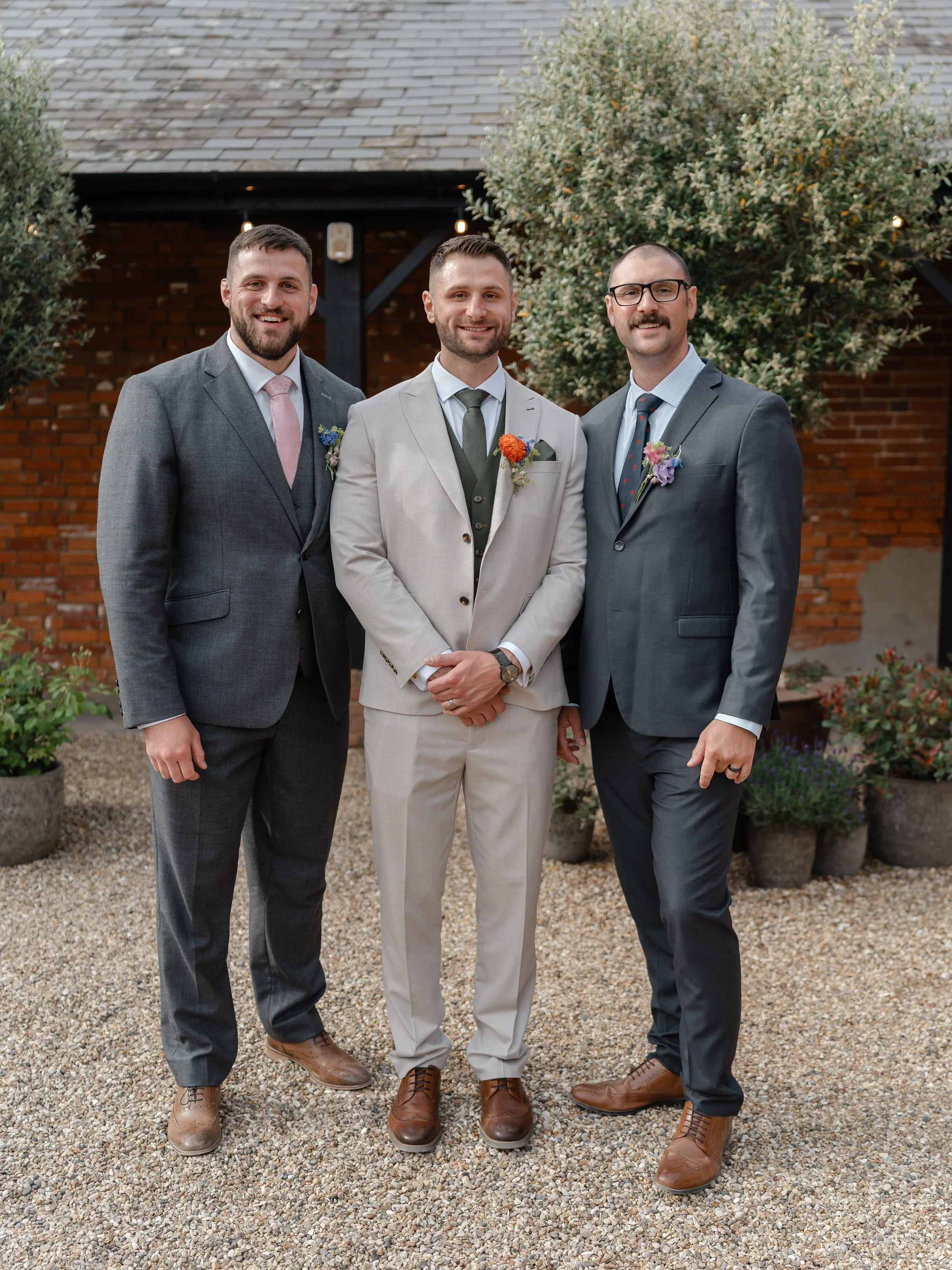 Three men dressed in suits, standing outside in front of brick building with trees, posing for a photo at a wedding or formal event.
