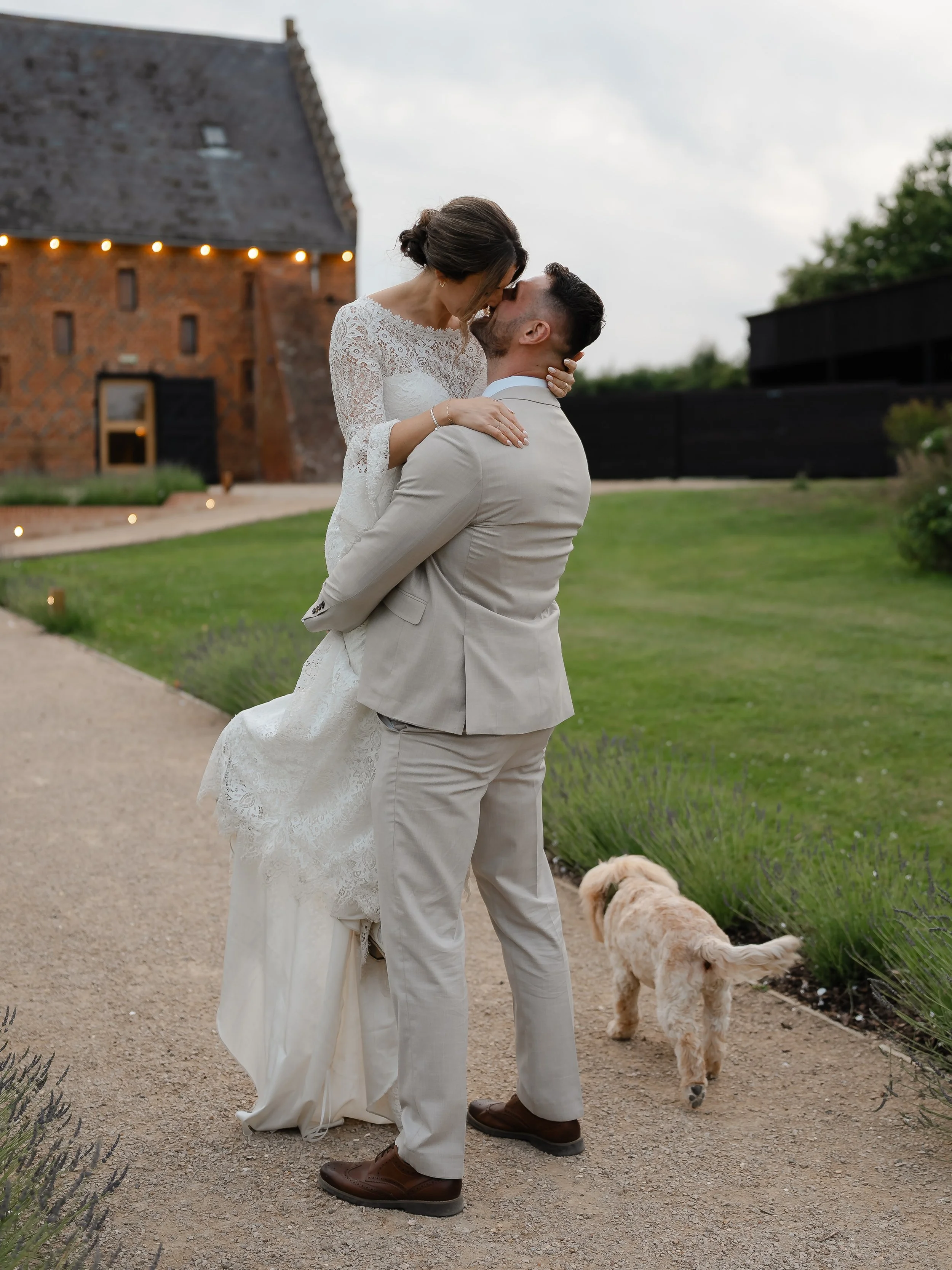 A man and woman dressed in wedding attire embrace and kiss outdoors, with the woman lifted in the man's arms. A small dog walks nearby, and a brick building with string lights is in the background.