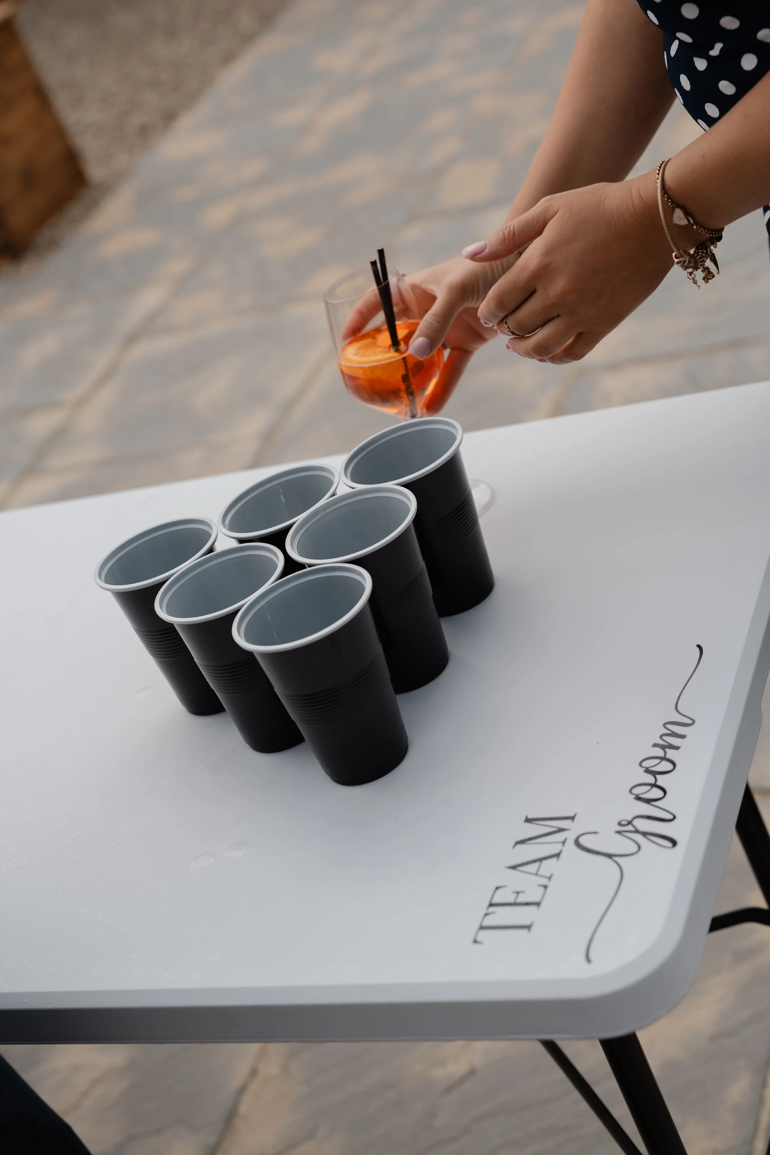A person placing an orange-colored drink with a slice of orange and two black straws into a black disposable cup on a white table with multiple other black cups