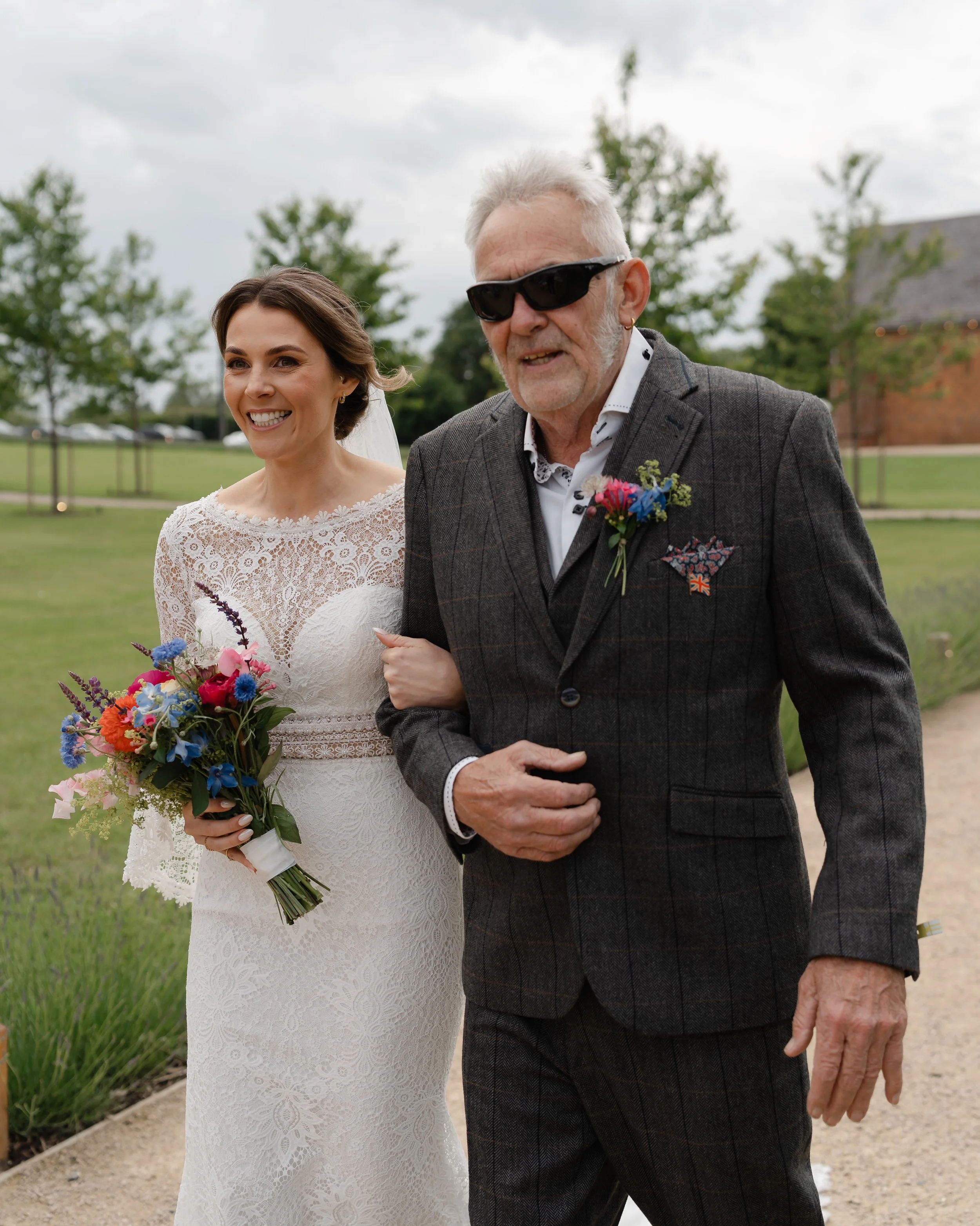 A bride in a white lace wedding dress holding a colorful bouquet, walking arm-in-arm with an older man dressed in a dark gray suit with a boutonniere, outdoors on a cloudy day with green trees and a building in the background.