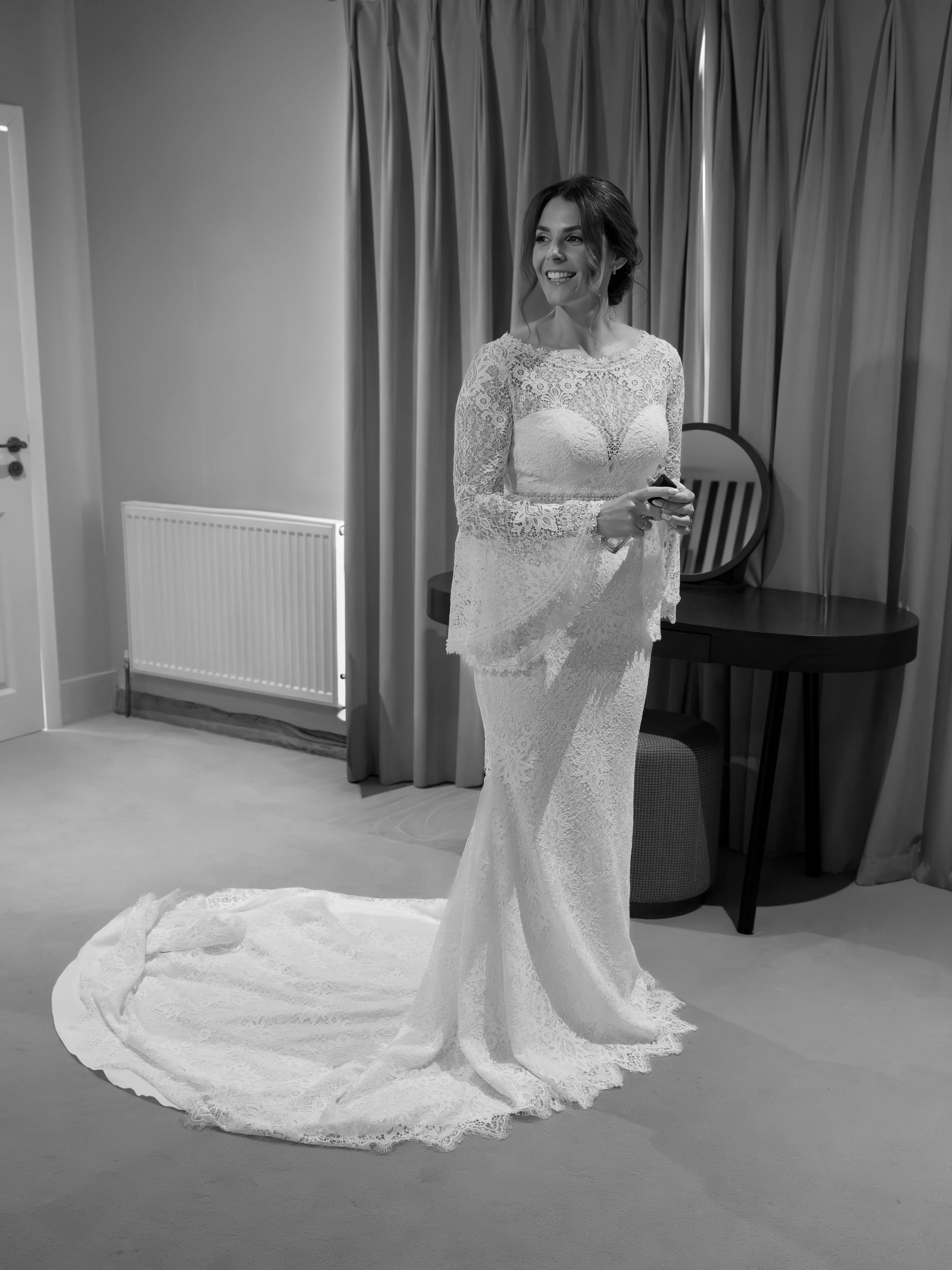 A smiling woman in a long lace wedding dress holding a phone, standing in a room with curtains and chairs, among wedding decor.
