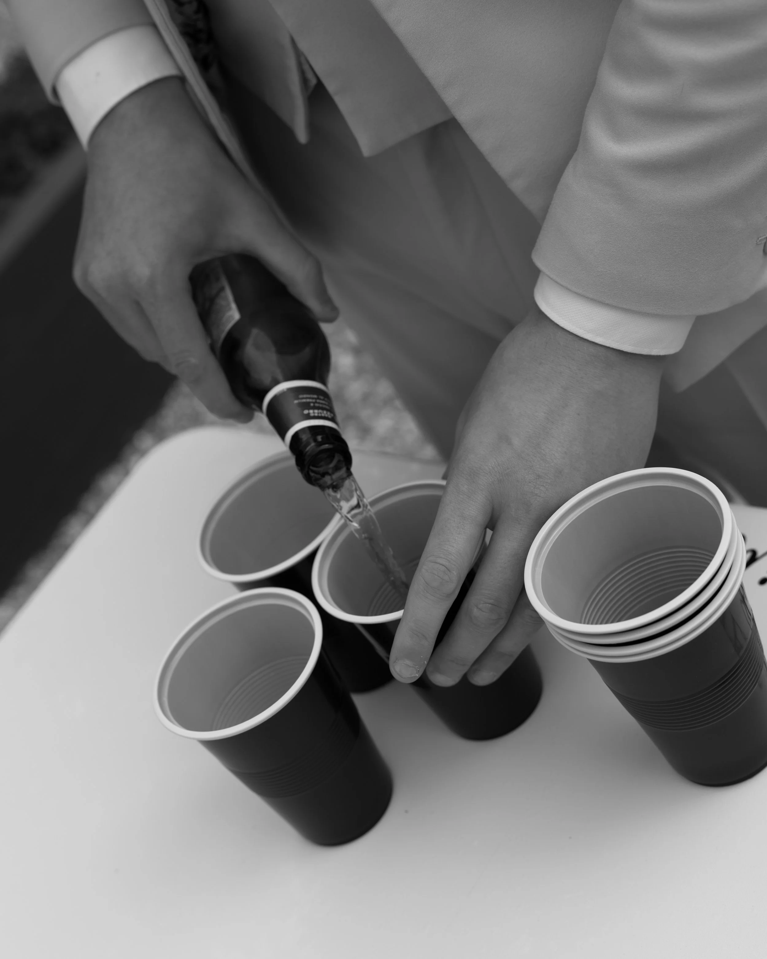 A person pouring dark liquid from a bottle into a white disposable cup, with several similar cups lined up on a surface.