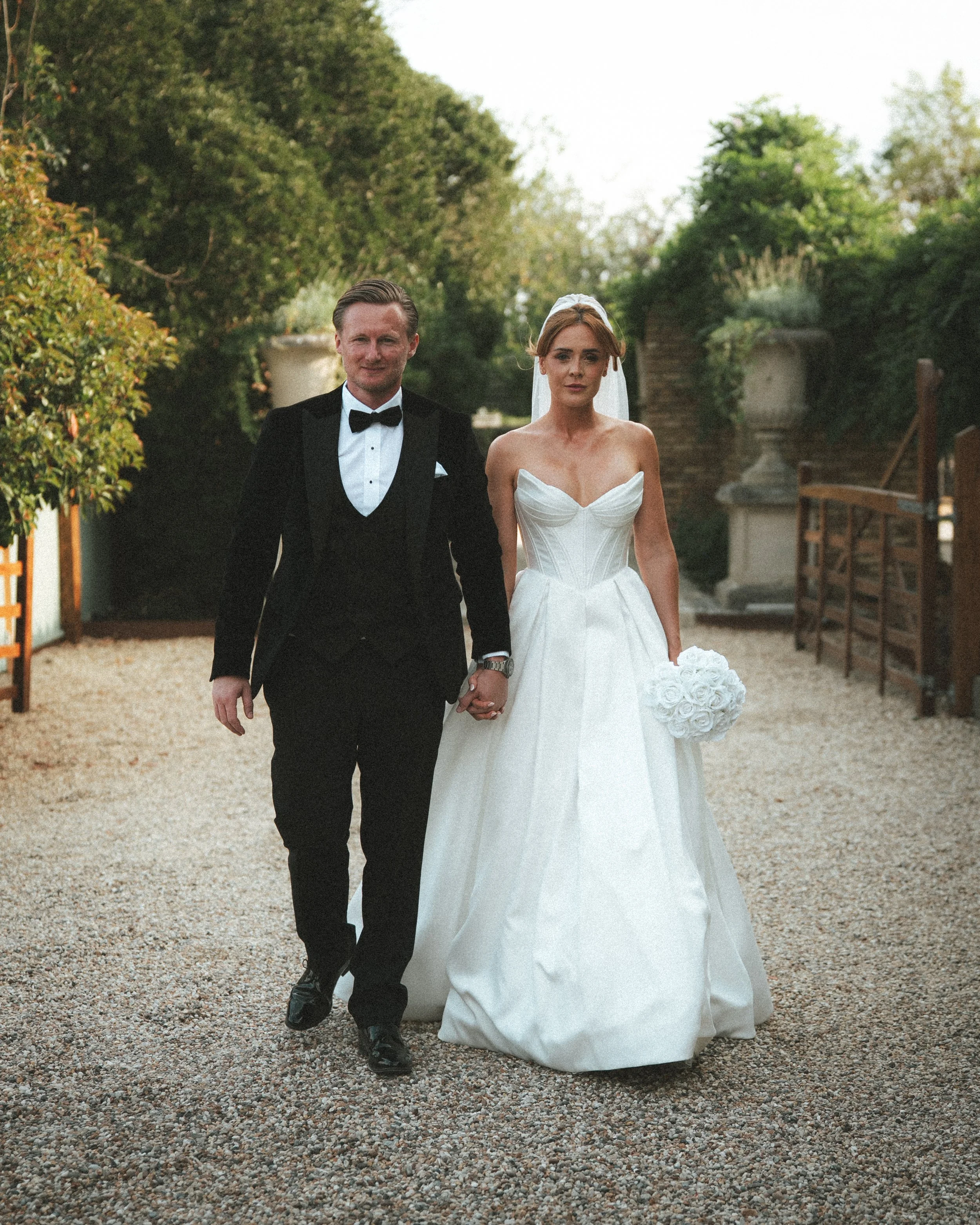A newlywed couple walking hand in hand outdoors: the groom in a black tuxedo with a bow tie, the bride in a strapless white wedding gown holding a bouquet of white roses, both smiling as they walk on a gravel path surrounded by greenery.
