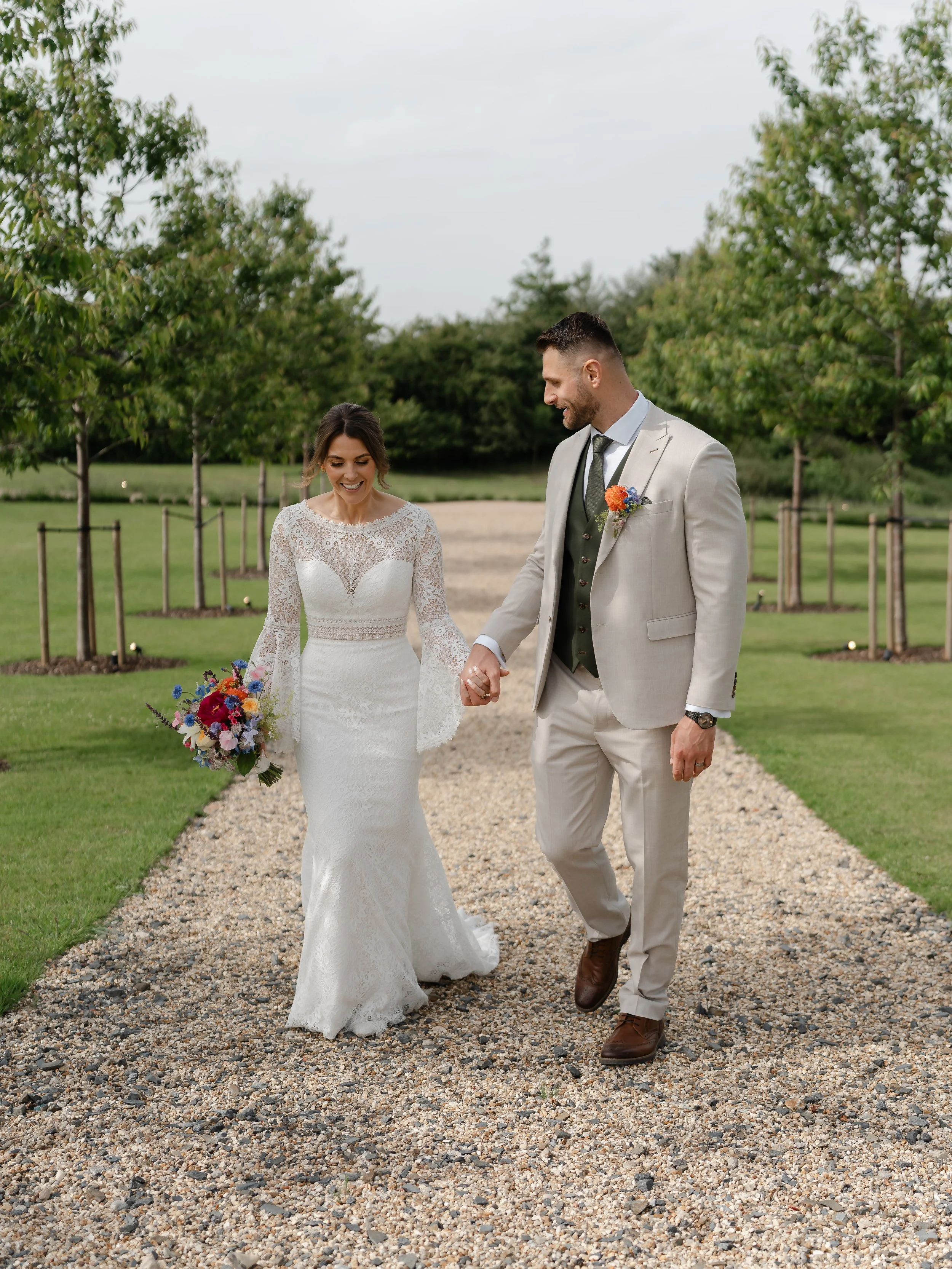 A bride and groom walking hand in hand outdoors on a gravel path, surrounded by green trees, during their wedding day. The bride is wearing a lace wedding dress and holding a bouquet, and the groom is dressed in a light-colored suit with a boutonnier