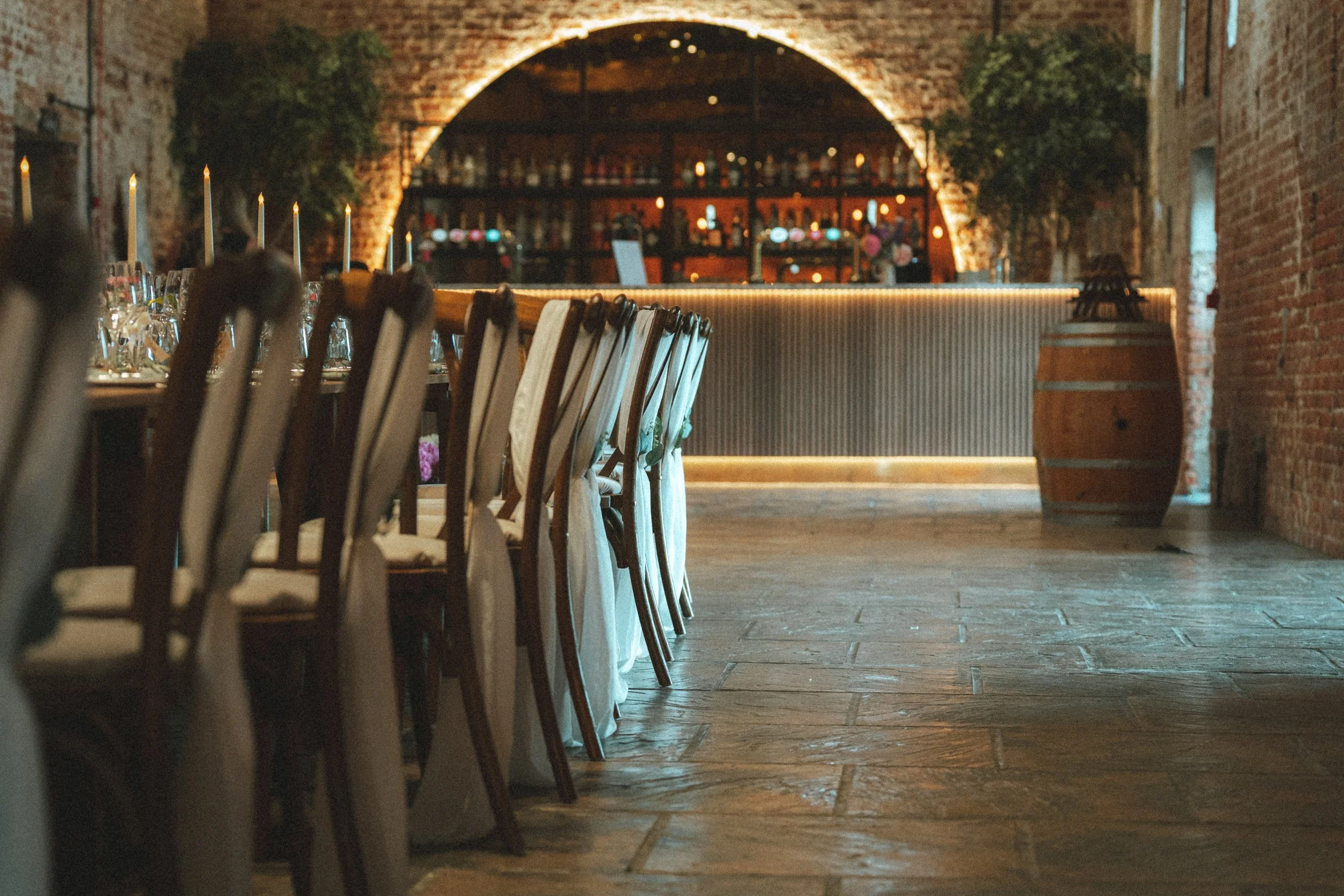 Empty restaurant dining area with rows of chairs, tables set with wine glasses and candles, exposed brick walls, and a bar in the background with lighting.